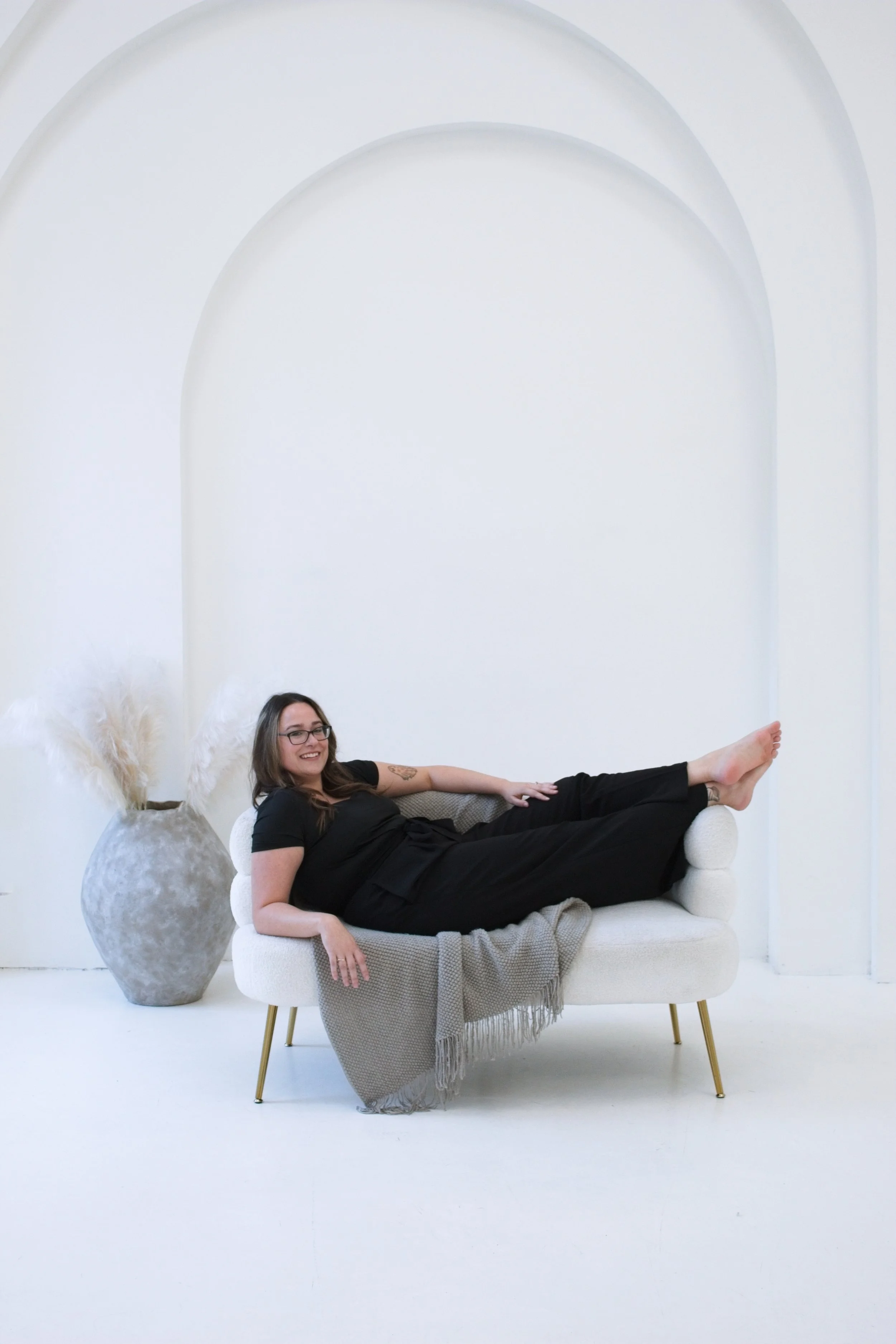 A woman with glasses and a black outfit relaxing on a white sofa in a minimalist, white room, with a large grey vase with pampas grass beside her.