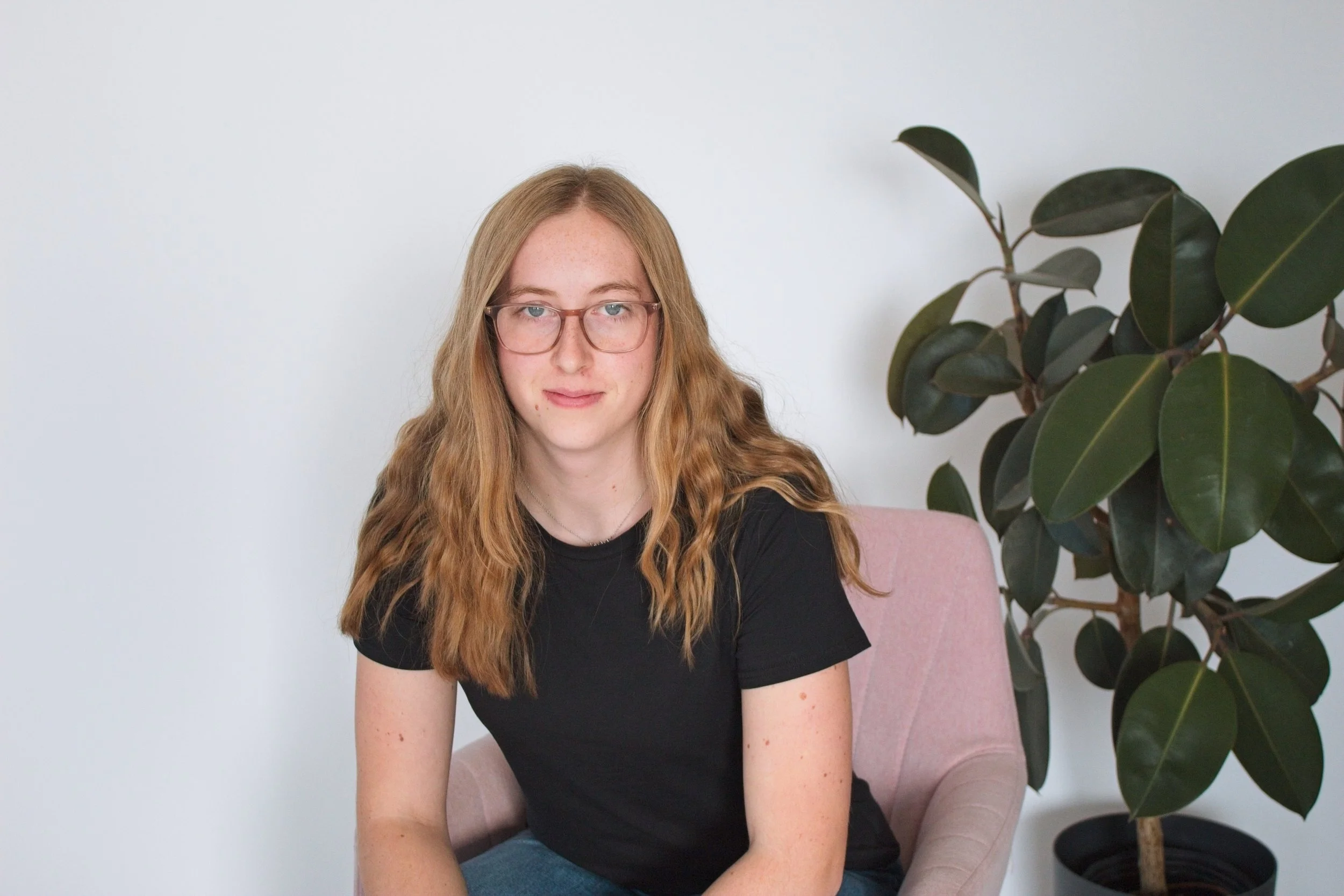A young woman with long wavy red hair, glasses, and fair skin sitting on a pink chair next to a large indoor plant with dark green leaves, against a plain white wall.