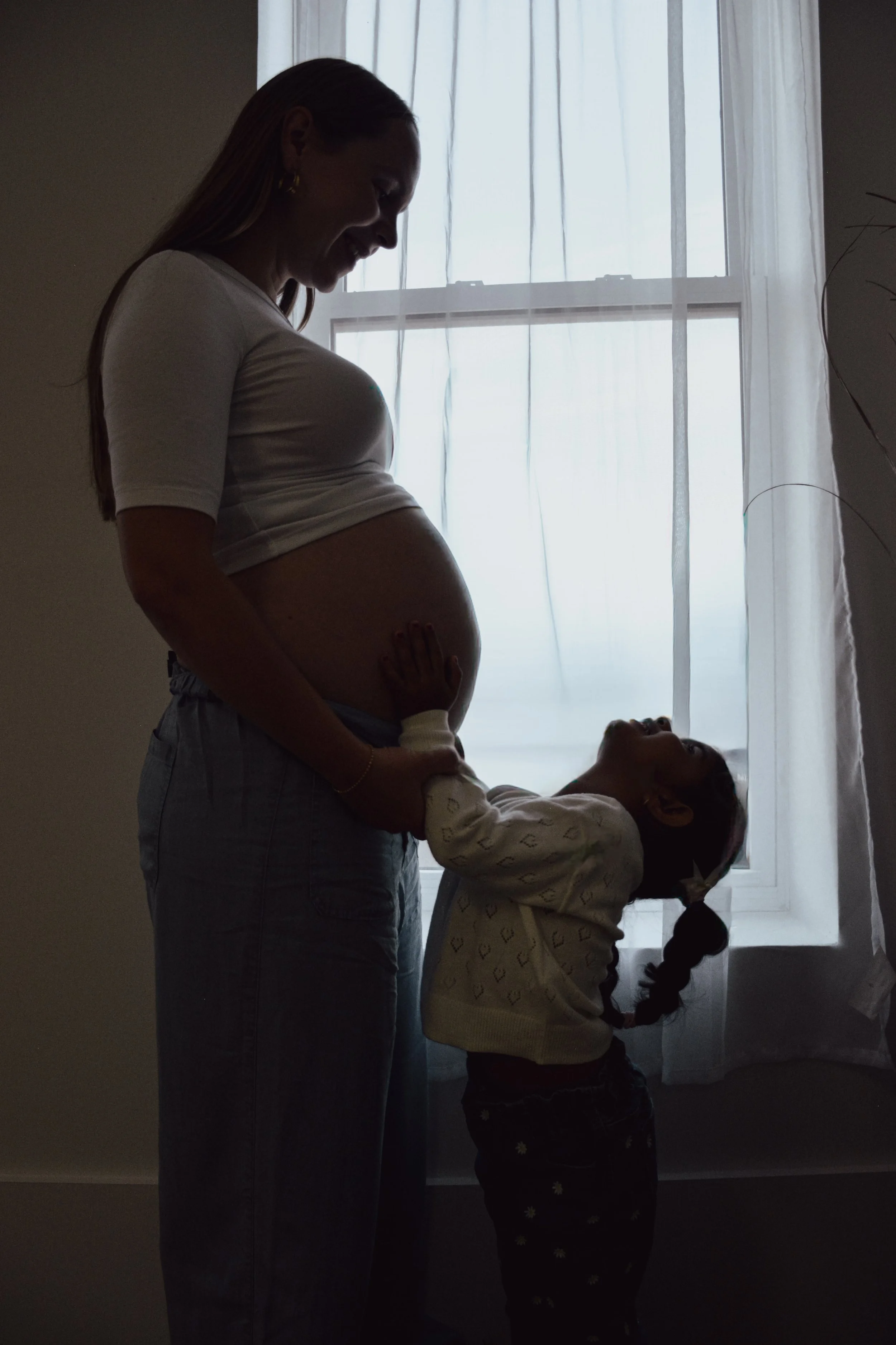 A pregnant woman smiling and touching her belly, standing near a window, with a young girl hugging her from behind.
