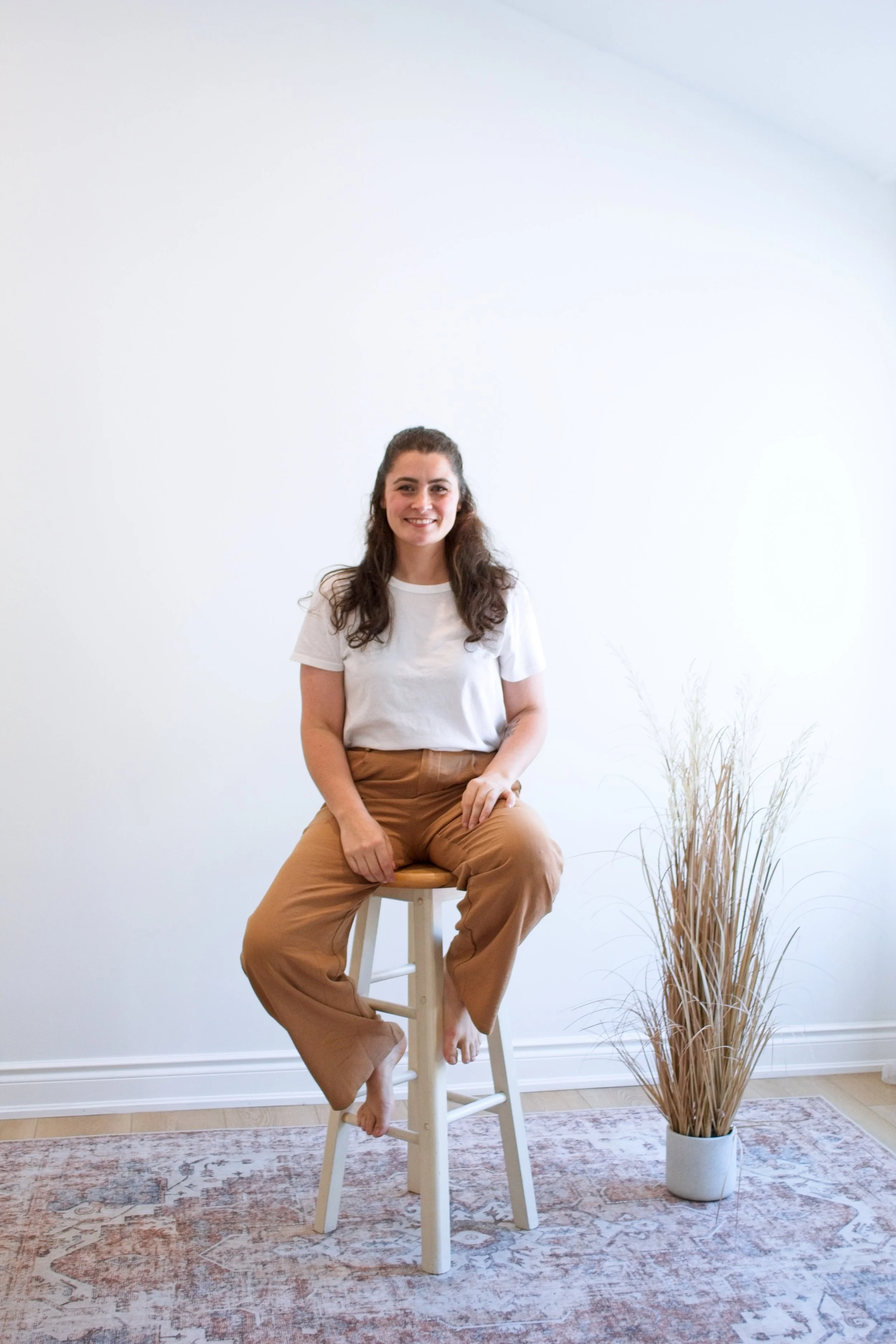 A woman with long dark hair, sitting on a wooden stool with one foot resting on the rung, smiling at the camera. She is wearing a white t-shirt and brown pants, with a white vase of dried grass to her right on a patterned rug against a plain white wall.