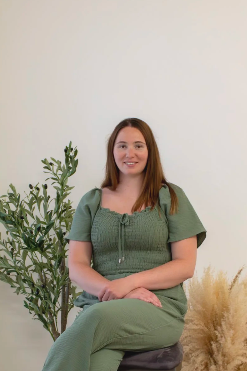 A woman with long brown hair sitting on a stool, wearing a green outfit, next to a green plant and some dried pampas grass, against a plain white wall background.