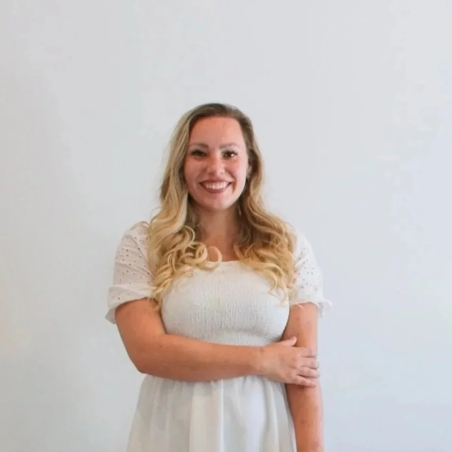 A woman with long blonde hair wearing a white dress smiling and standing with her arms crossed against a plain white wall