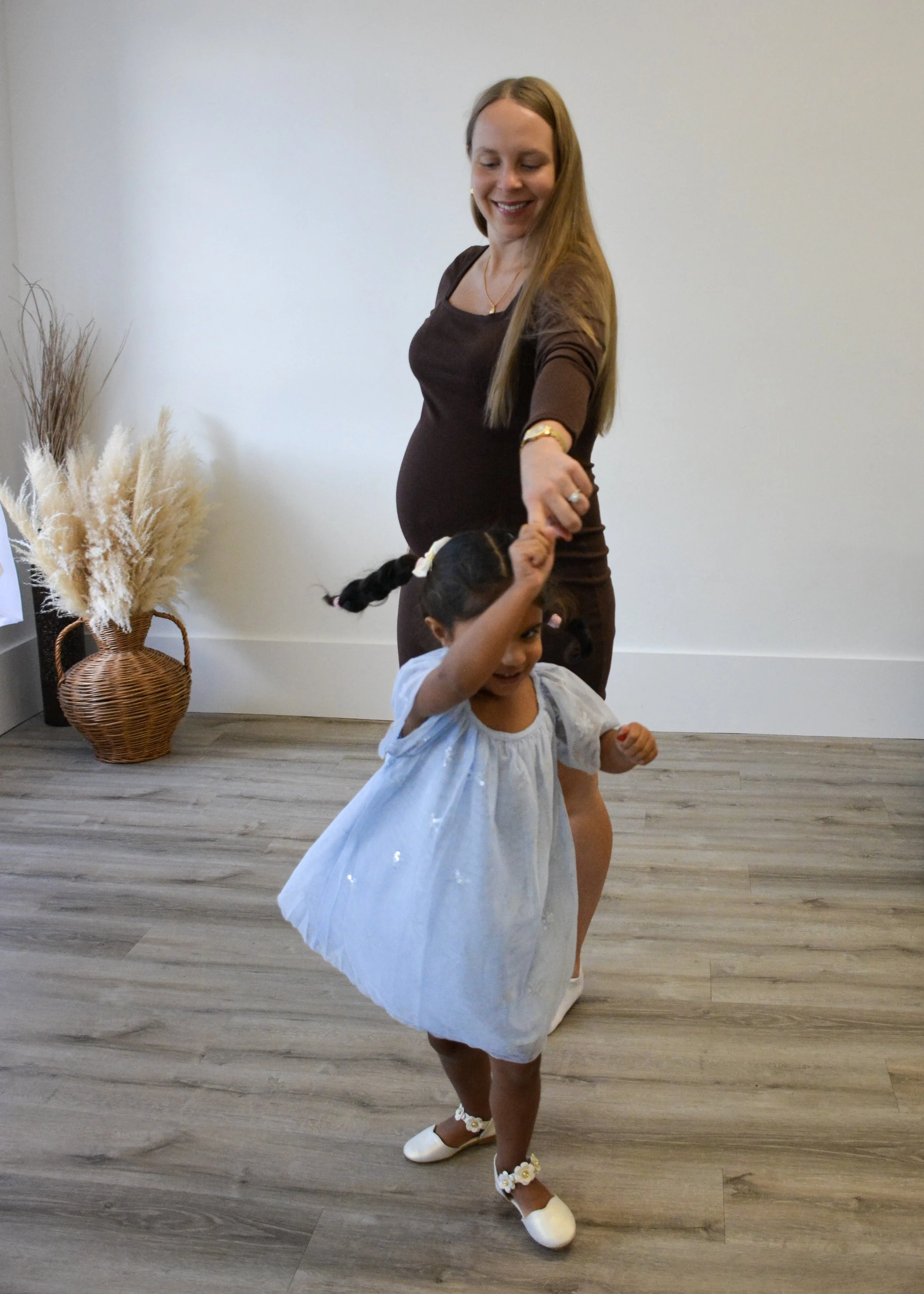A pregnant woman and young girl dancing together indoors. The woman is wearing a brown dress and the girl is dressed in a light blue dress with white shoes and floral accessories. They are smiling and holding hands, with the woman twirling the girl.