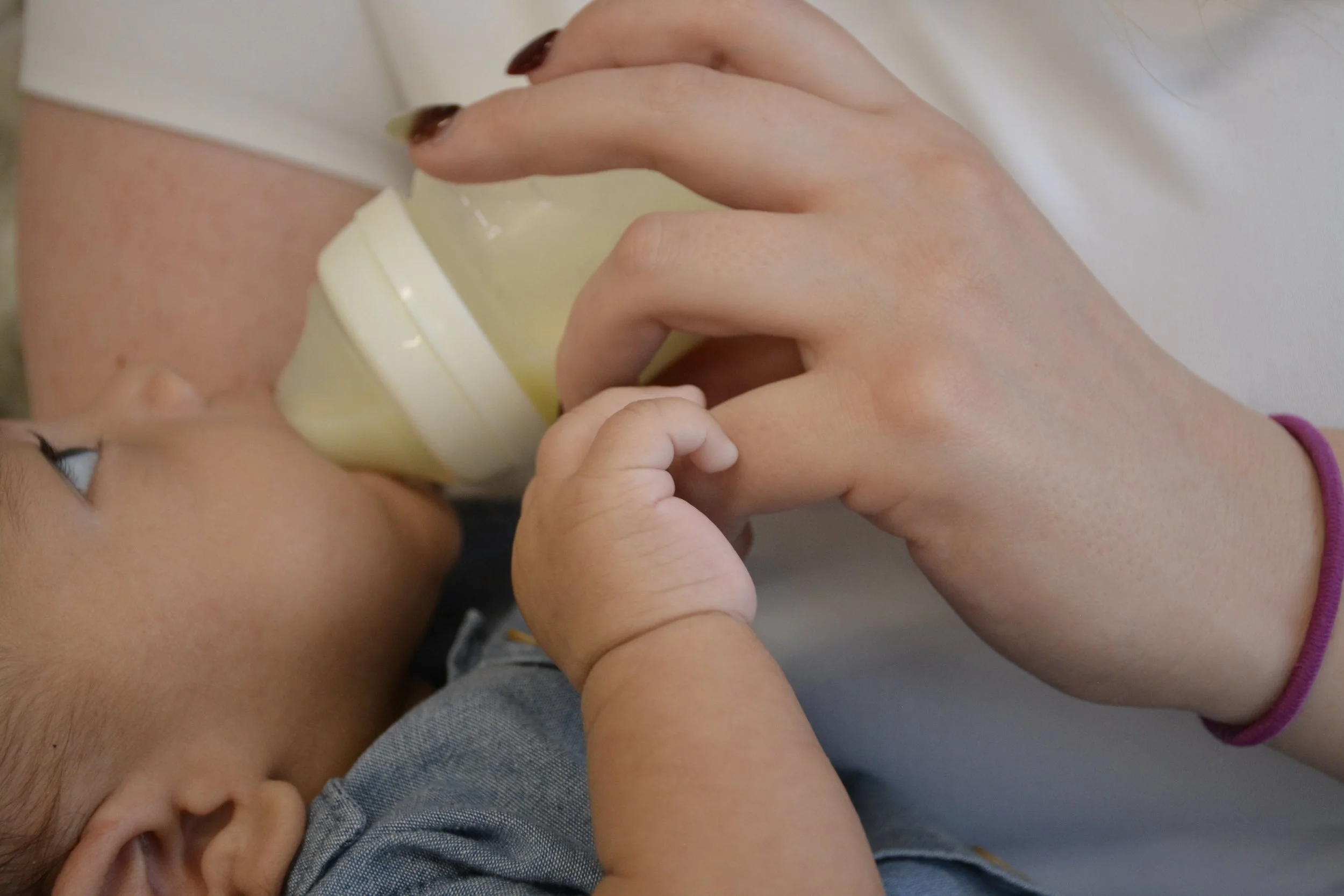 A young child being fed with a bottle, held by an adult hand. The child's eyes are looking up at the bottle. The adult is wearing a purple hair tie on their wrist.