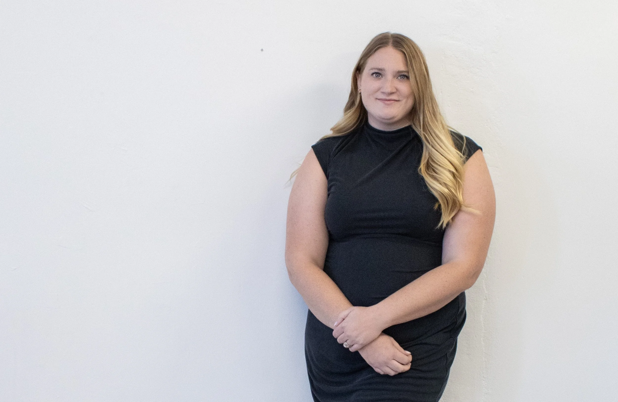 A woman with long blonde hair, wearing a black sleeveless dress, standing against a plain white wall with her hands clasped in front of her.