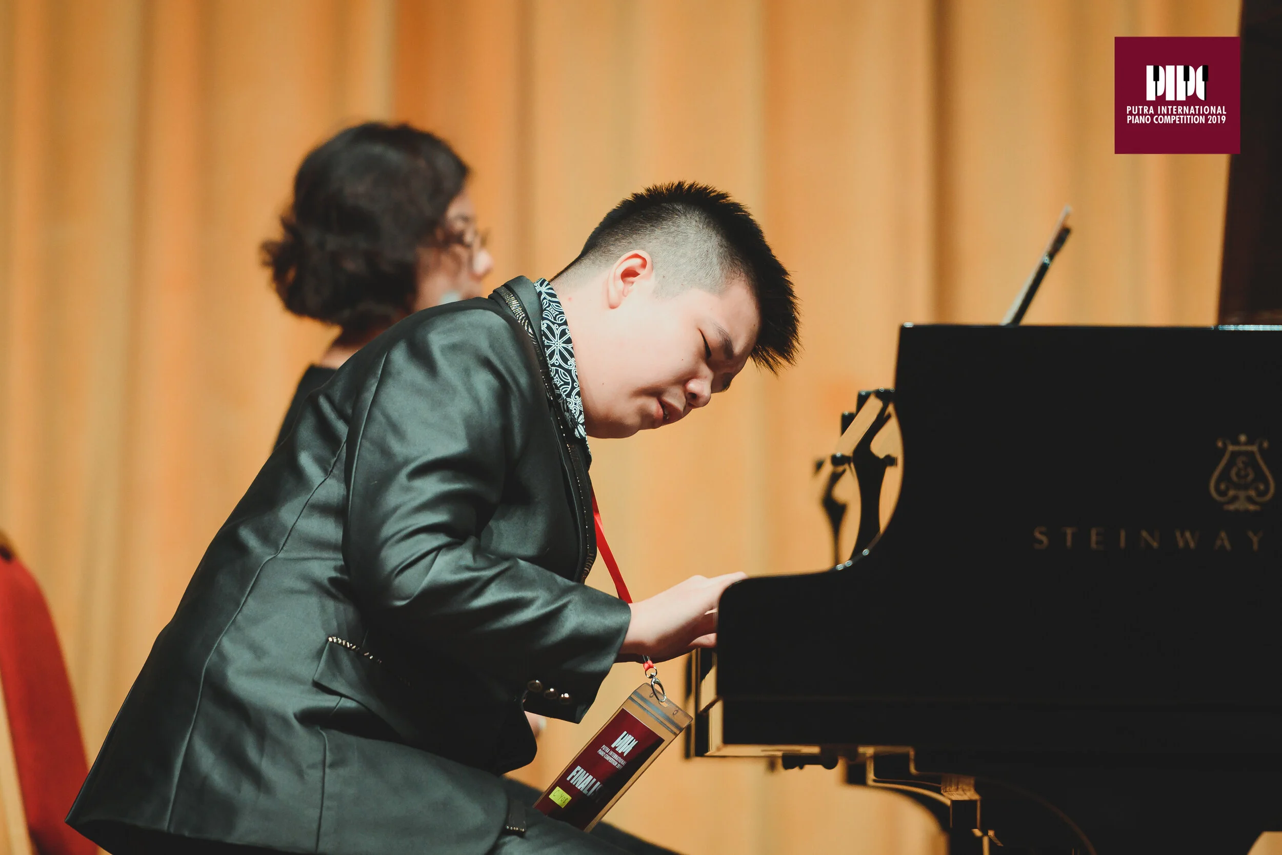 Pianist playing a Steinway piano at a competition, wearing a dark blazer and a badge, with a blurred audience in the background.
