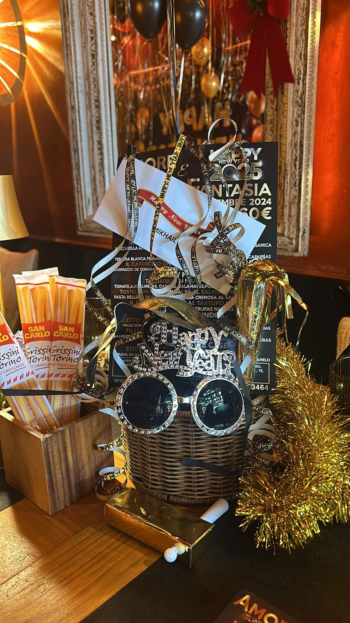 Decoración de celebración de Año Nuevo en un restaurante, con un cartel que dice 'Feliz Año Nuevo' y otros textos, un espejo con globos dorados y negros, una cesta con gafas negras decoradas con cristales, y guirnaldas doradas.