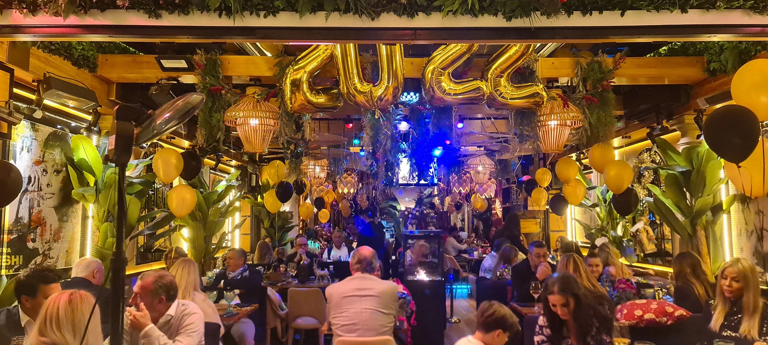 Interior de restaurante decorado con globos amarillos y negros, plantas grandes, luces decorativas, y globos con letras doradas que formaron la palabra 'GULA', con muchas personas comiendo y socializando.