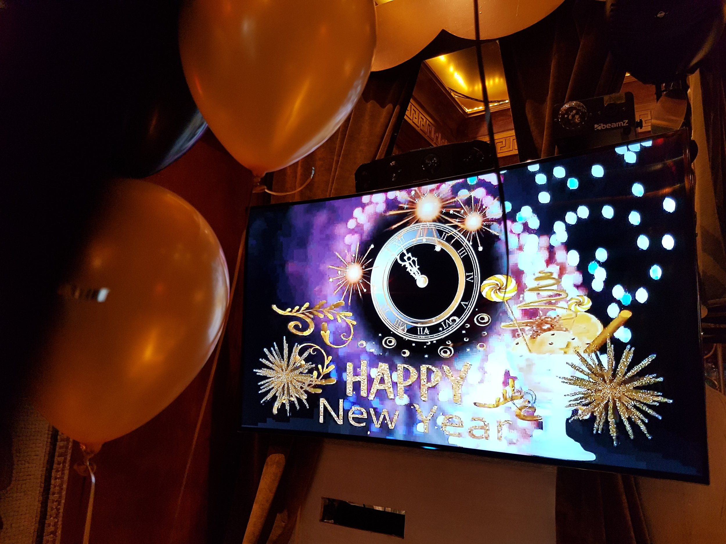 Pantalla con reloj, fuegos artificiales y mensaje de feliz año nuevo, decoraciones doradas y globos en un ambiente festivo.
