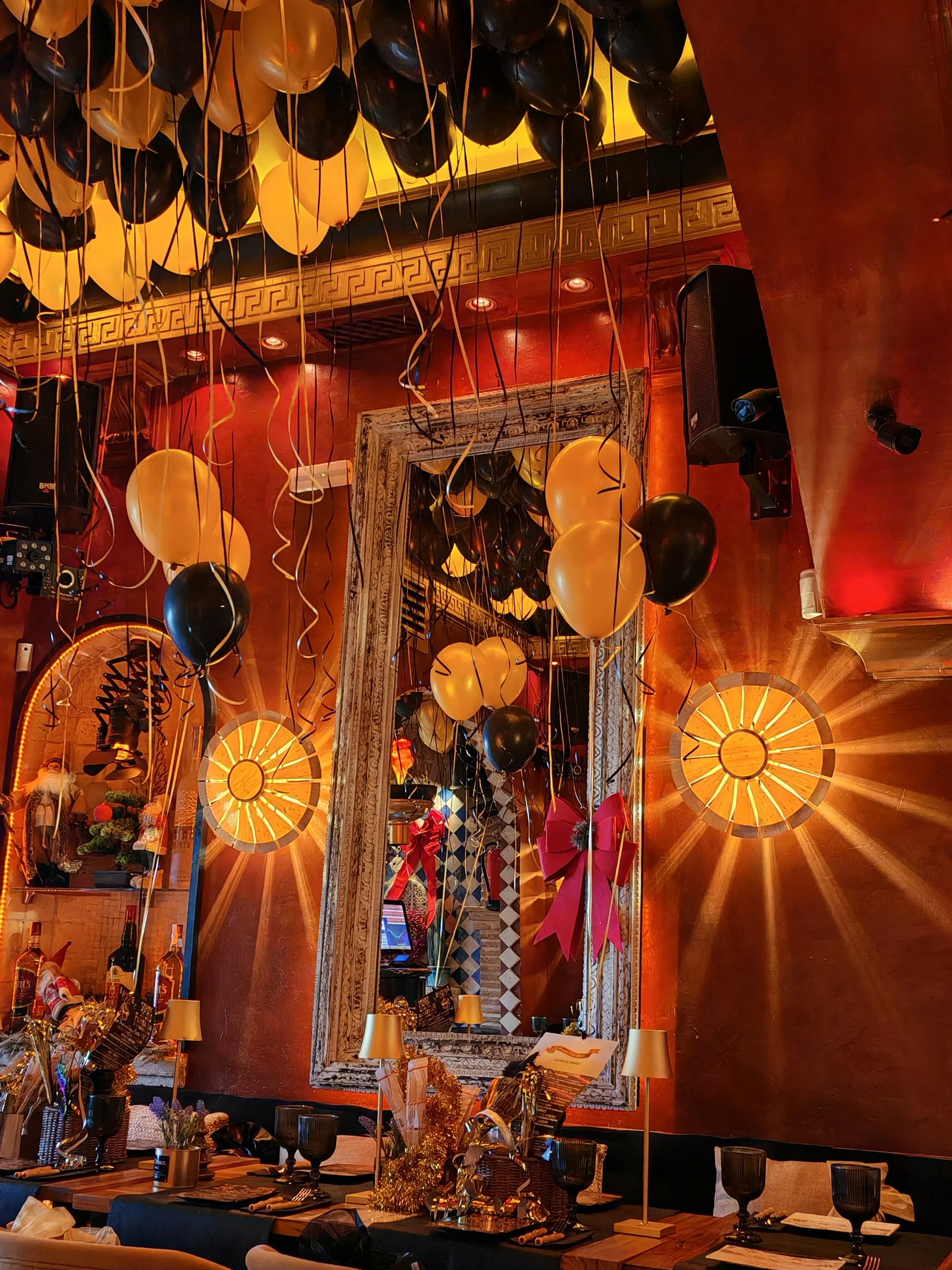 Decoración de una celebración con globos negros y dorados, un gran espejo decorativo, mesas con decoraciones festivas y platos, y una iluminación cálida en un restaurante o salón.