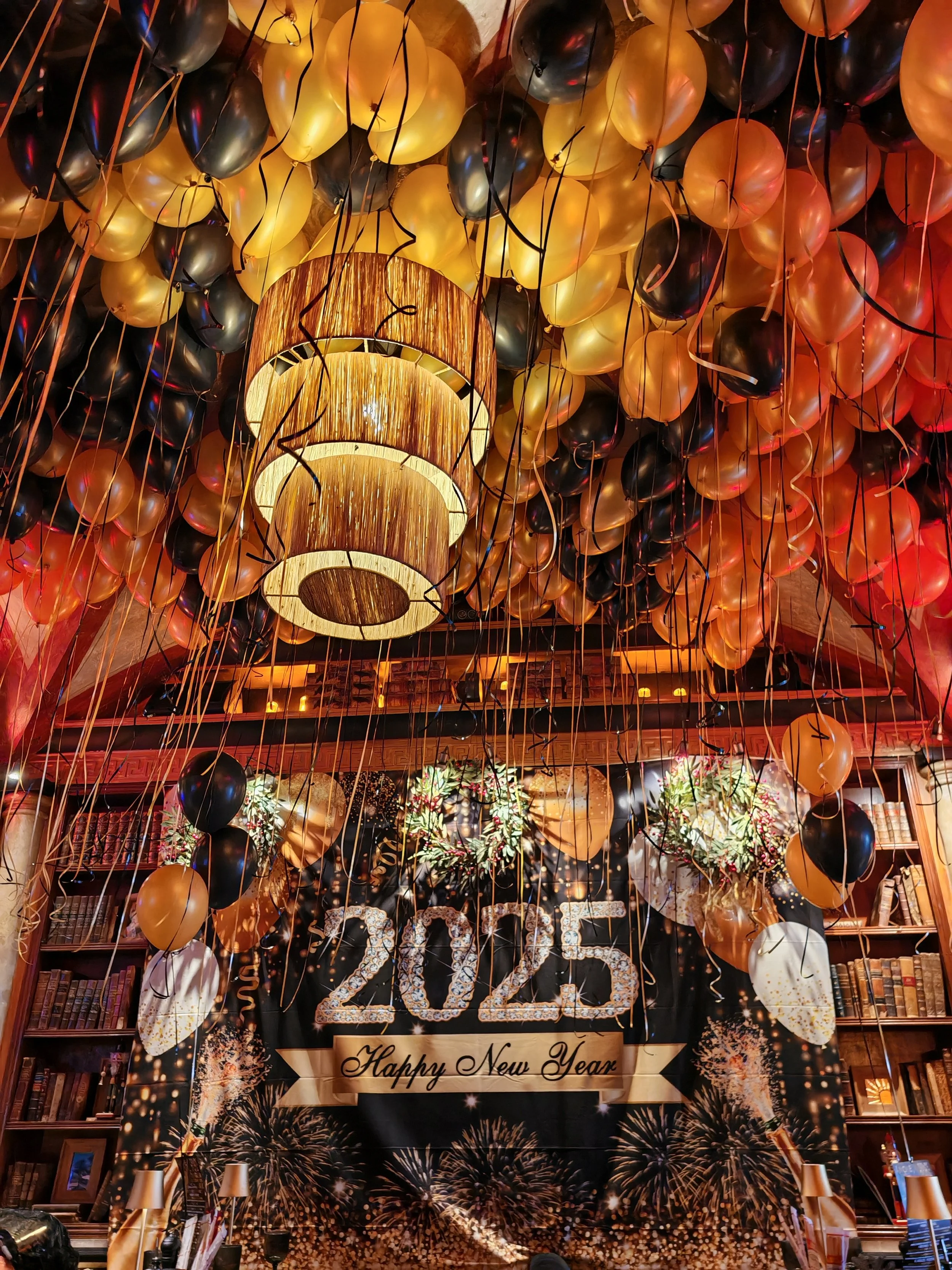 Decoraciones de celebración de Año Nuevo 2025 con globos dorados, negros y plateados en el techo y un cartel que dice 'Feliz Año Nuevo' y '2025' con fuegos artificiales.