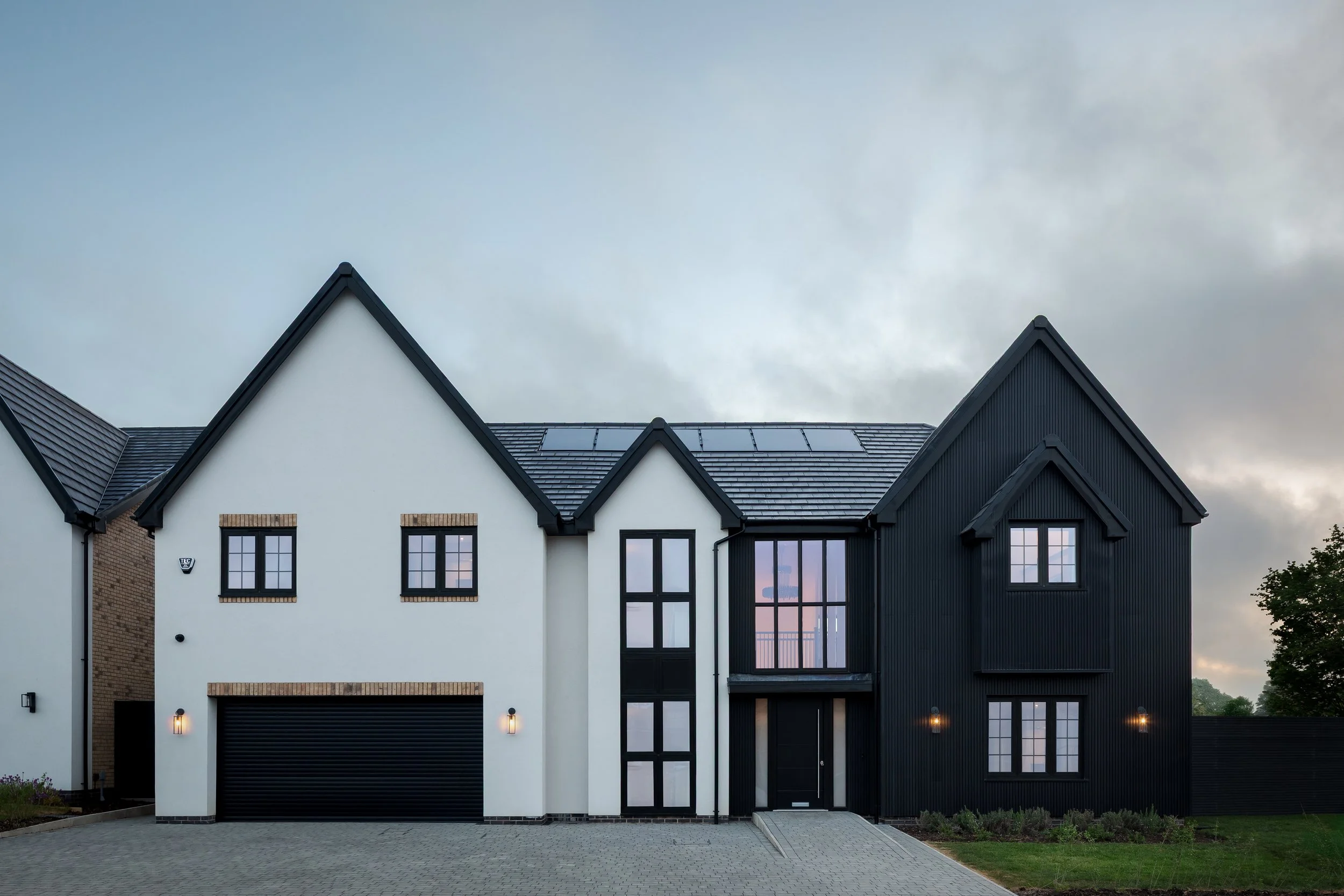 A modern two-story house with a white and black exterior, gabled roof, large windows, and a closed garage door, set against a cloudy sky.