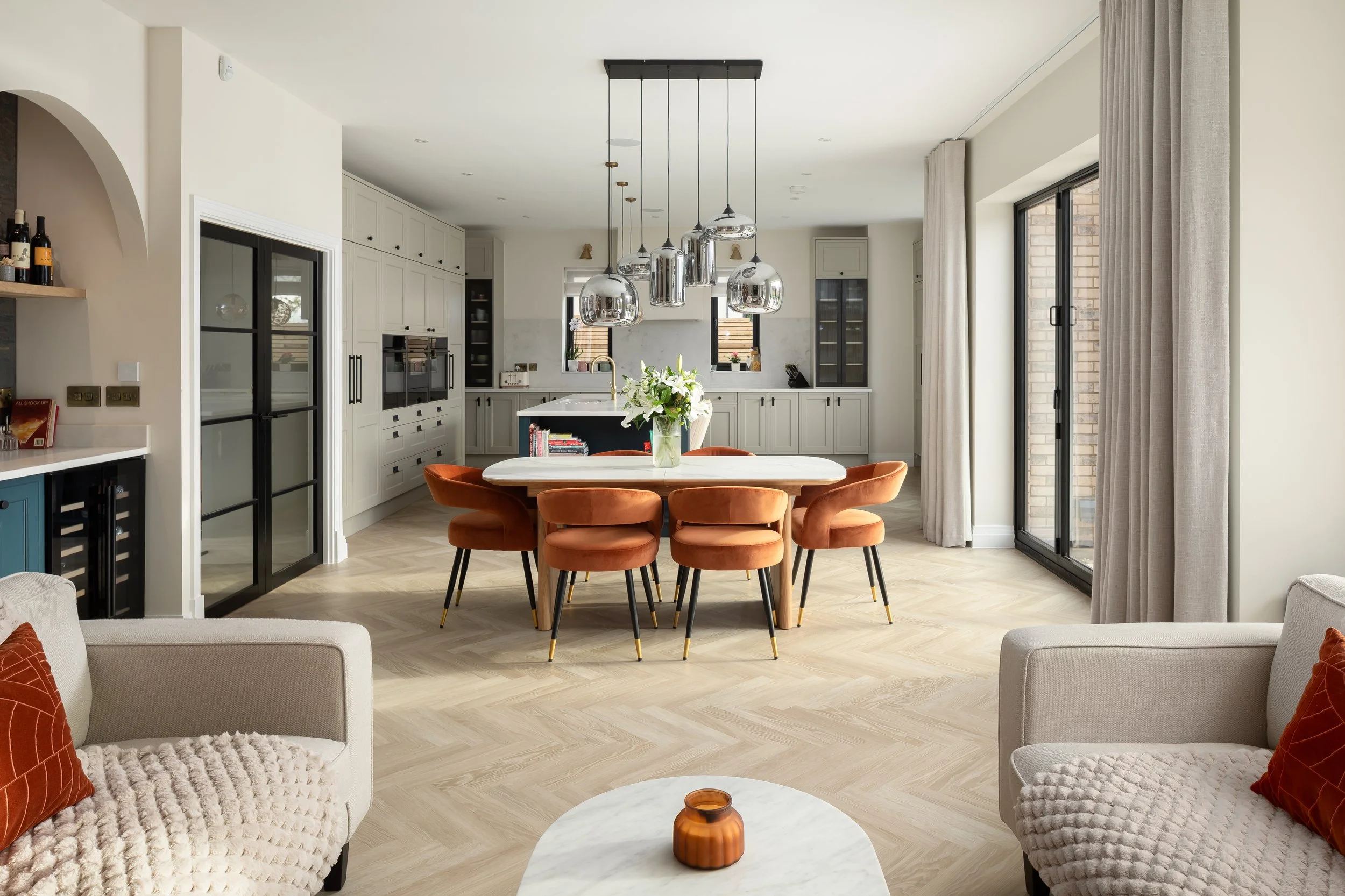 Modern open-concept kitchen and dining area with a round table, six orange velvet chairs, and a pendant light fixture, with a living space including beige armchairs and a marble coffee table.