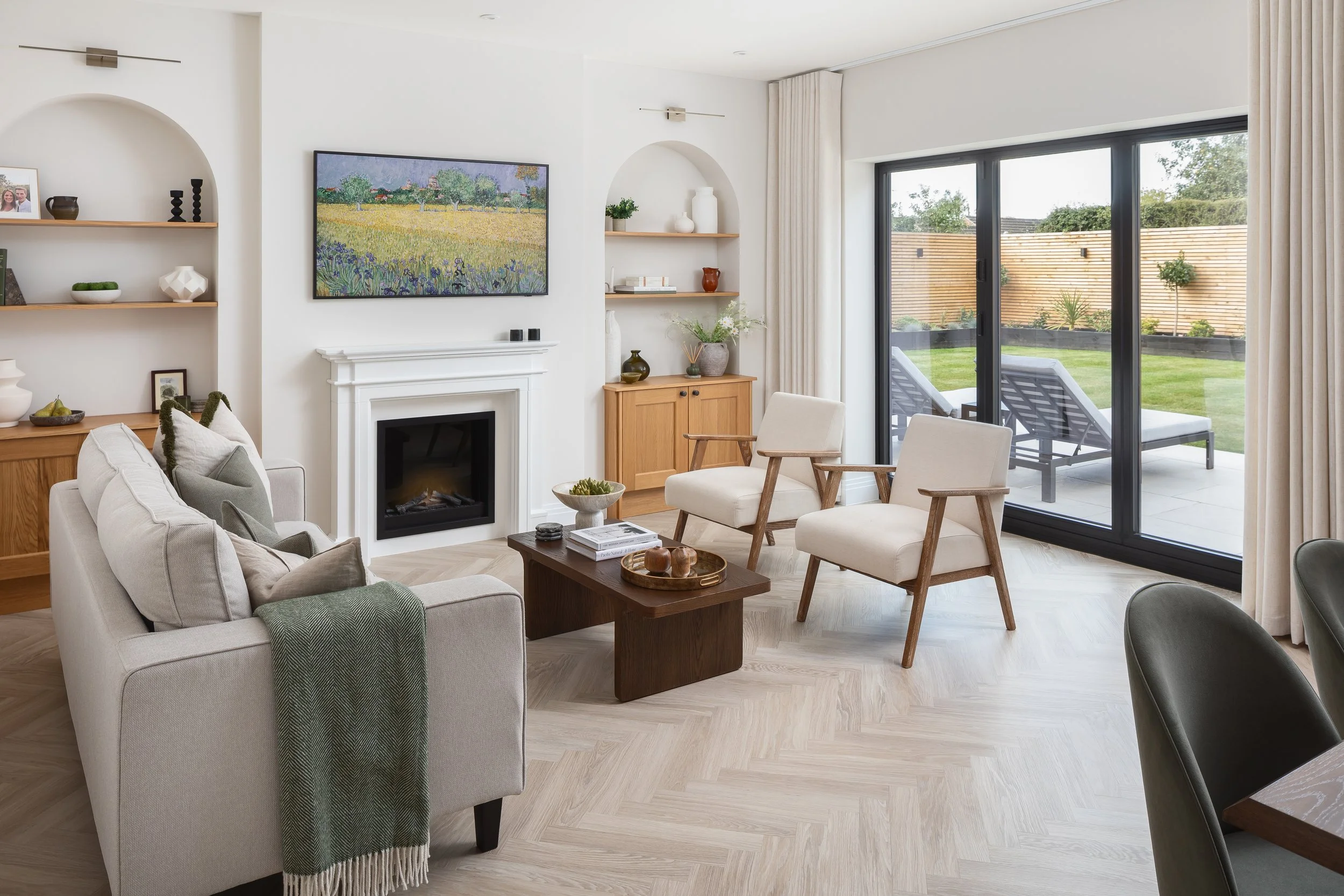 Modern living room with white walls, a fireplace, beige sofa, two armchairs, a coffee table, built-in shelves, and large glass sliding doors leading to a backyard patio with lounge chairs.
