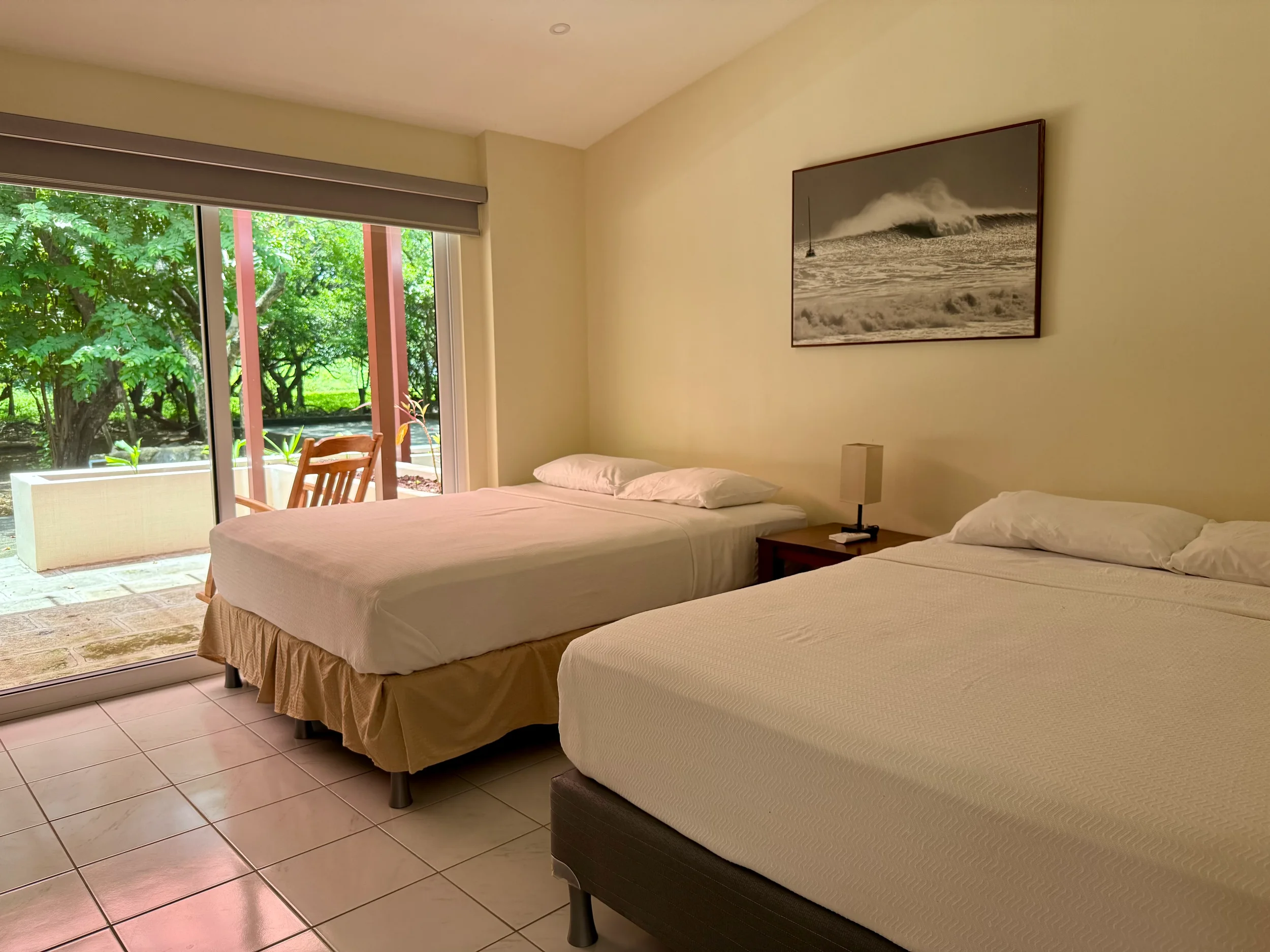A hotel room with two single beds, a nightstand with a lamp, a large window open to a patio with trees outside, and a black-and-white ocean-themed photograph on the wall.