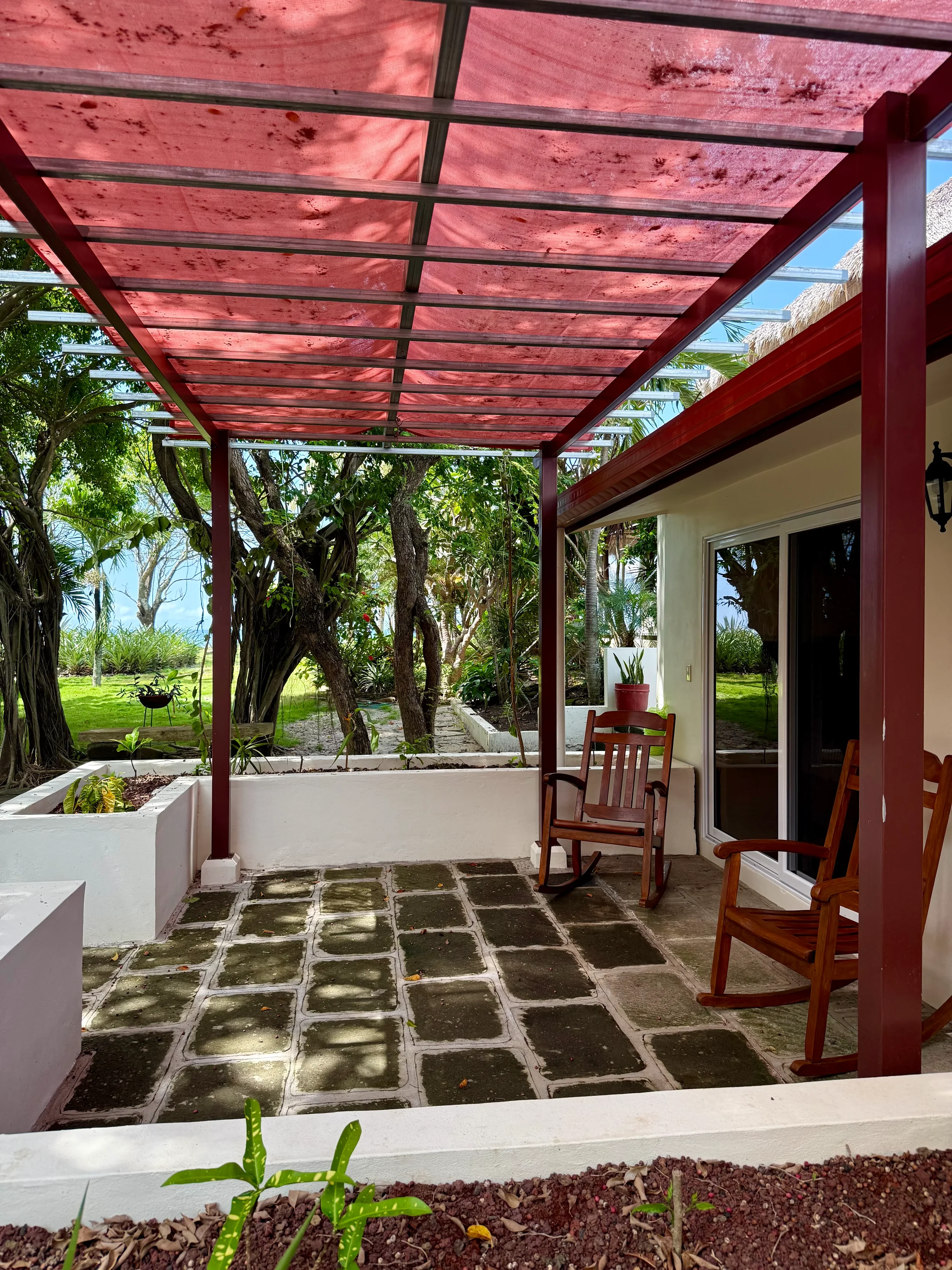 A covered patio with two wooden rocking chairs, a potted plant, and a garden with trees and grass in the background.