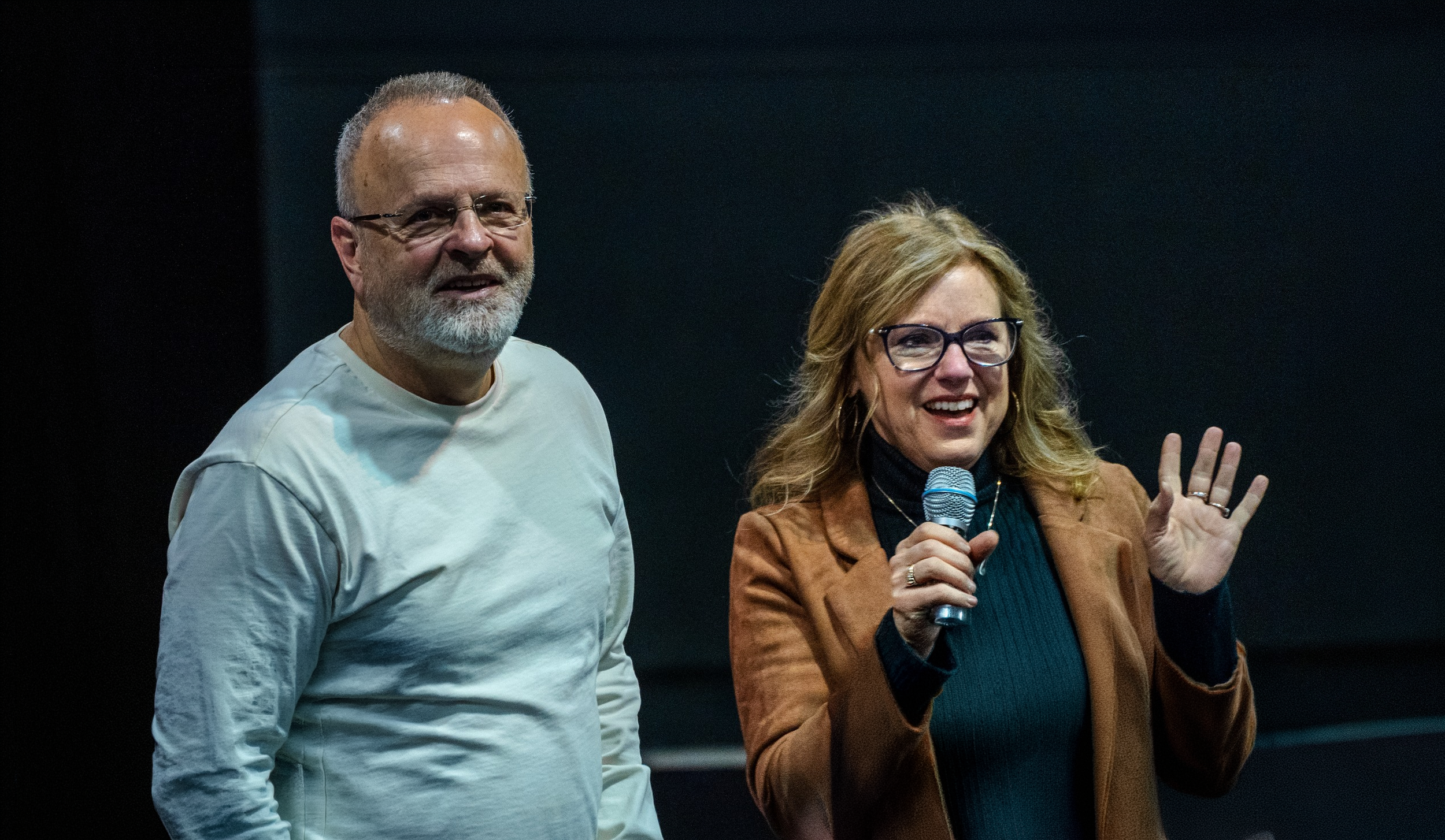 A man with glasses and a beard stands next to a woman with glasses and blond hair, who is holding a microphone and speaking, in a dark room.