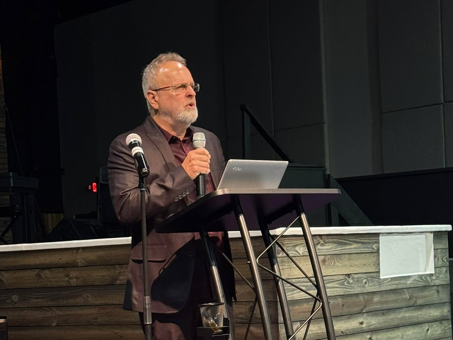 A man with gray hair and glasses, wearing a brown blazer, giving a speech at a podium with a laptop, microphone, and two additional microphones, in a dark, modern conference room.