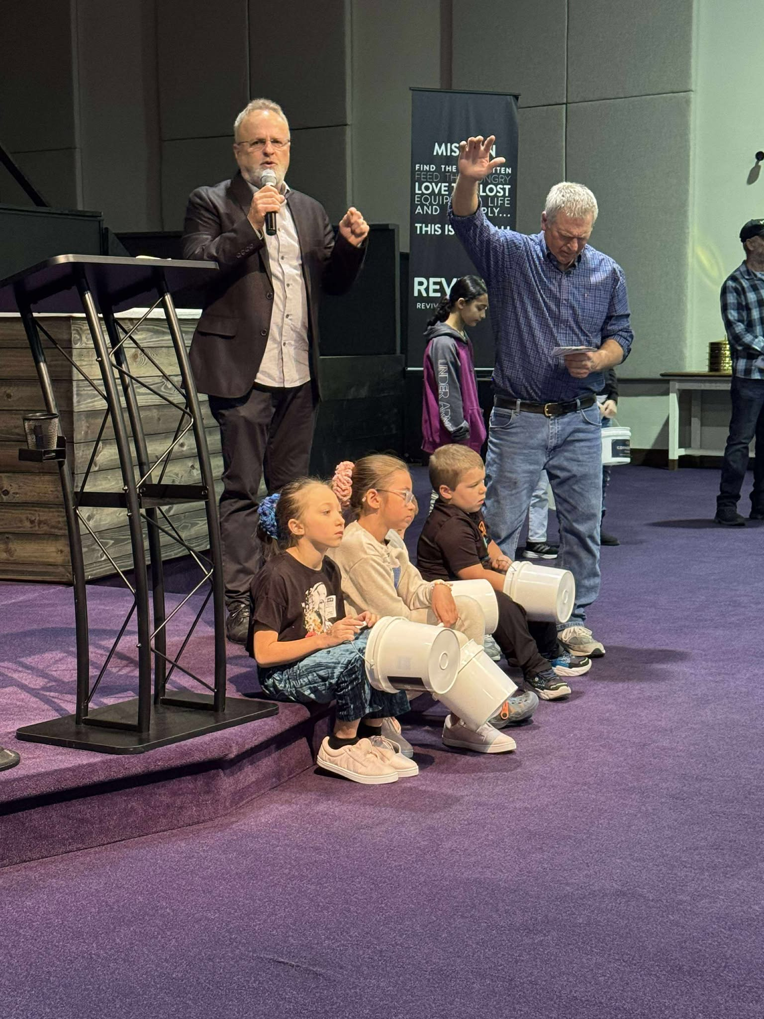 A man with glasses and a beard is speaking into a microphone on a stage, while another man with light hair and a checkered shirt is standing nearby holding a paper. Several children are sitting on the floor with white buckets in their laps, some wearing glasses and hair tied back, listening attentively. There is a banner behind the standing men with text that reads 'MISSION... FIND THE FOOD, FEED THE HUNGRY, LOVE THE LOST, EQUIP THE LIFE AND SUPPLY... THIS IS...' and a wooden podium on the stage.