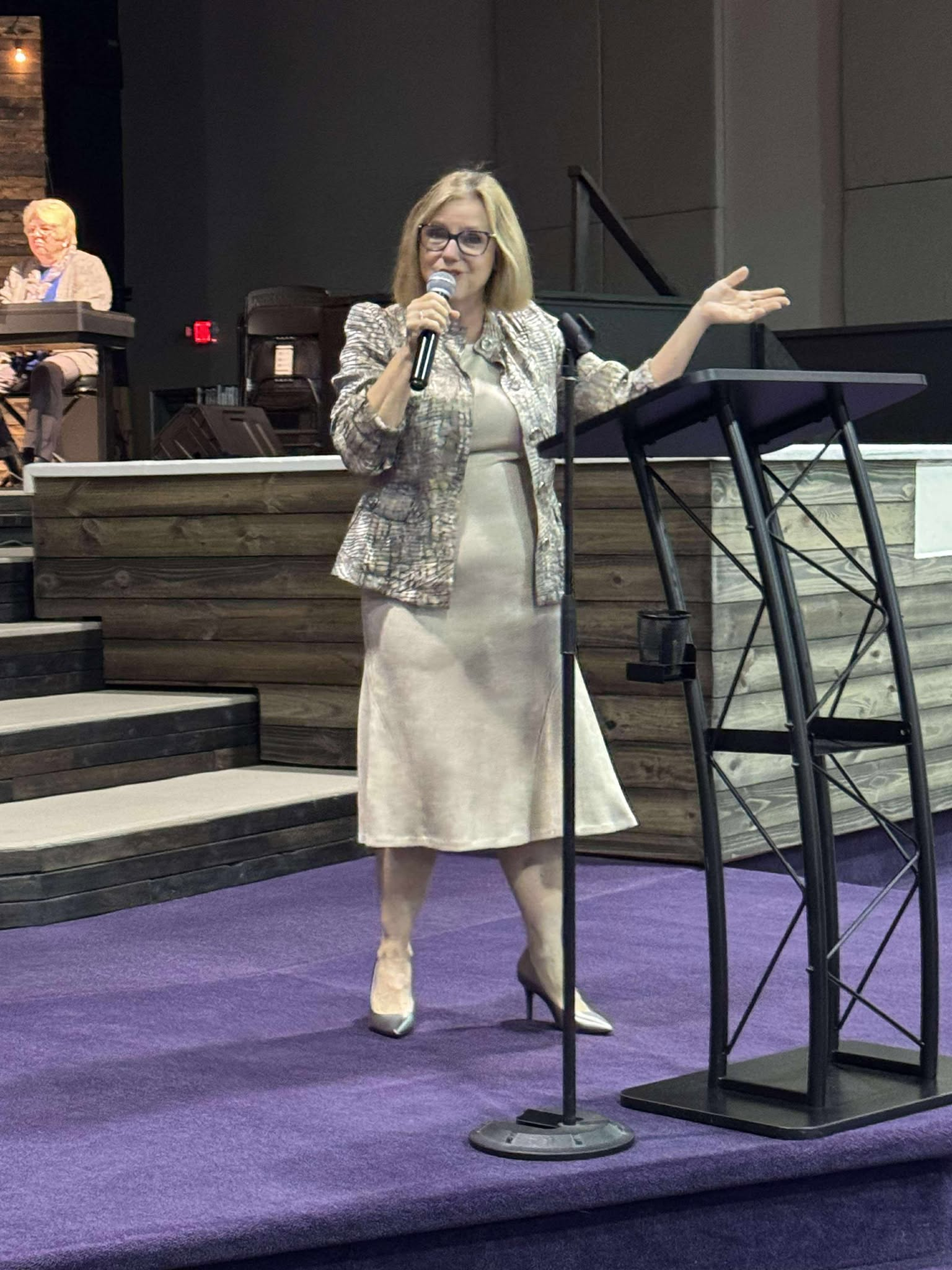 A woman with blonde hair and glasses is speaking into a microphone on a stage, gesturing with her right hand. She is wearing a patterned blazer, a cream-colored dress, and cream-colored boots. In the background, a woman with short hair, glasses, and a beige blazer is sitting at a table.