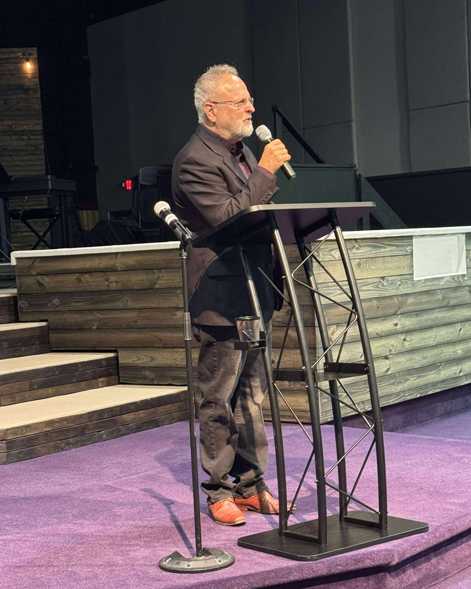 A man with glasses and gray hair speaking into a microphone at a podium on a stage with purple carpet and wooden steps.