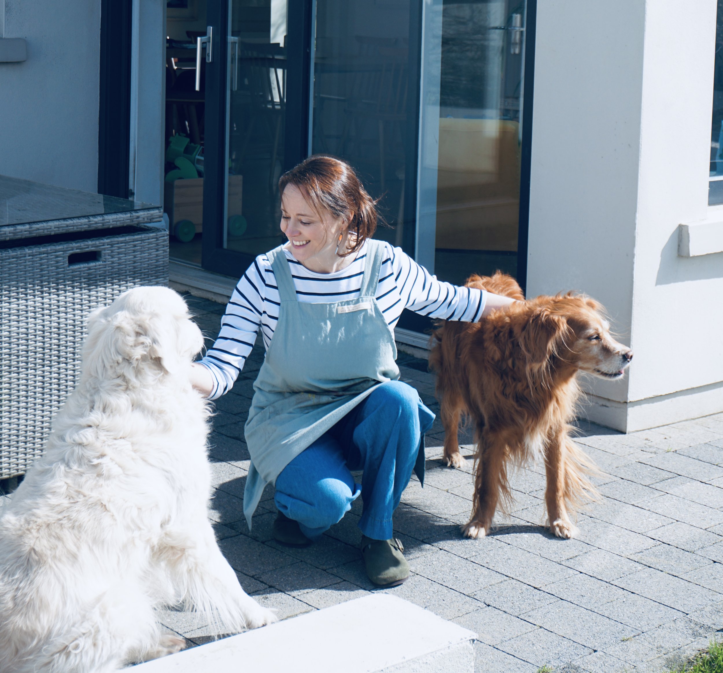 A woman with light brown hair, wearing a striped shirt and overalls, smiling and interacting with two dogs, a white fluffy one and a brown long-haired one, on a patio outside a house with sliding glass doors.
