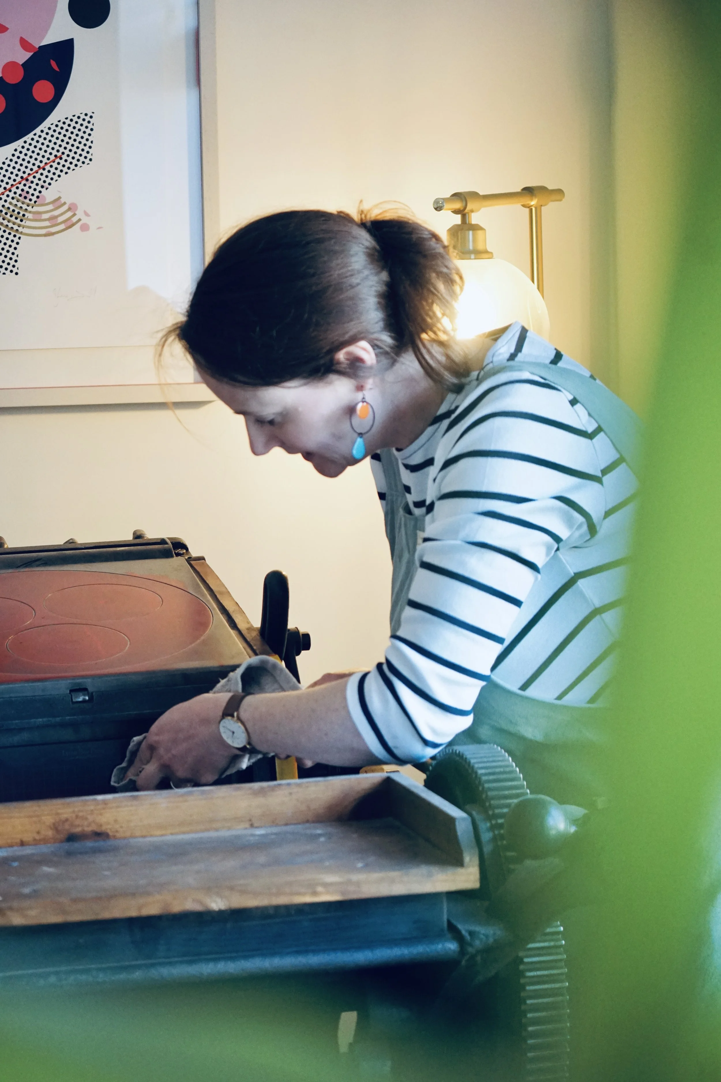 A woman with short brown hair, wearing a striped shirt and colorful earrings, is rolling dough at a wooden table in a cozy kitchen. A small tabletop pizza oven is on the counter, and a textured gear-shaped object is near the edge of the table. The background includes a yellow wall, a modern lamp, and an abstract art piece.