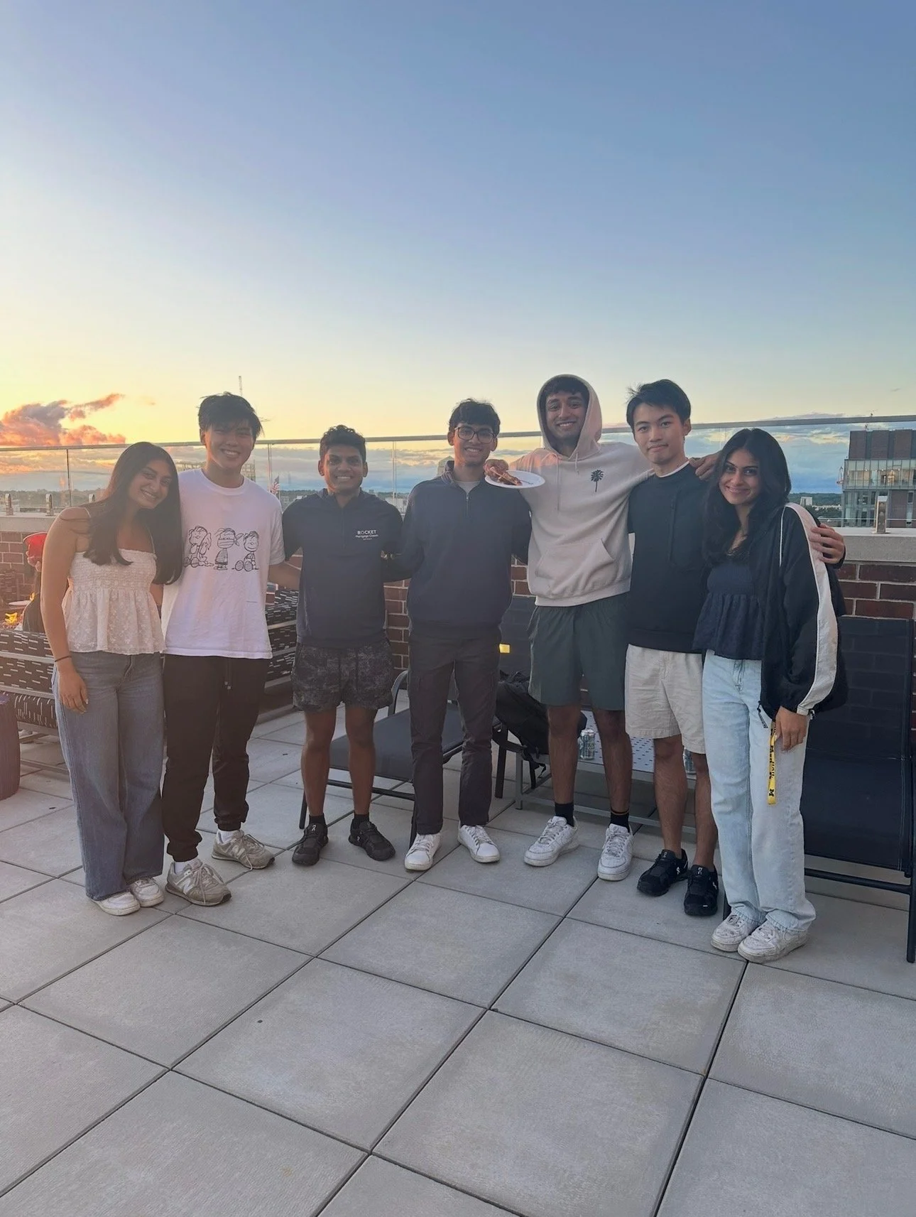 Group of eight young adults standing on a rooftop patio during sunset, smiling, with cityscape in the background.