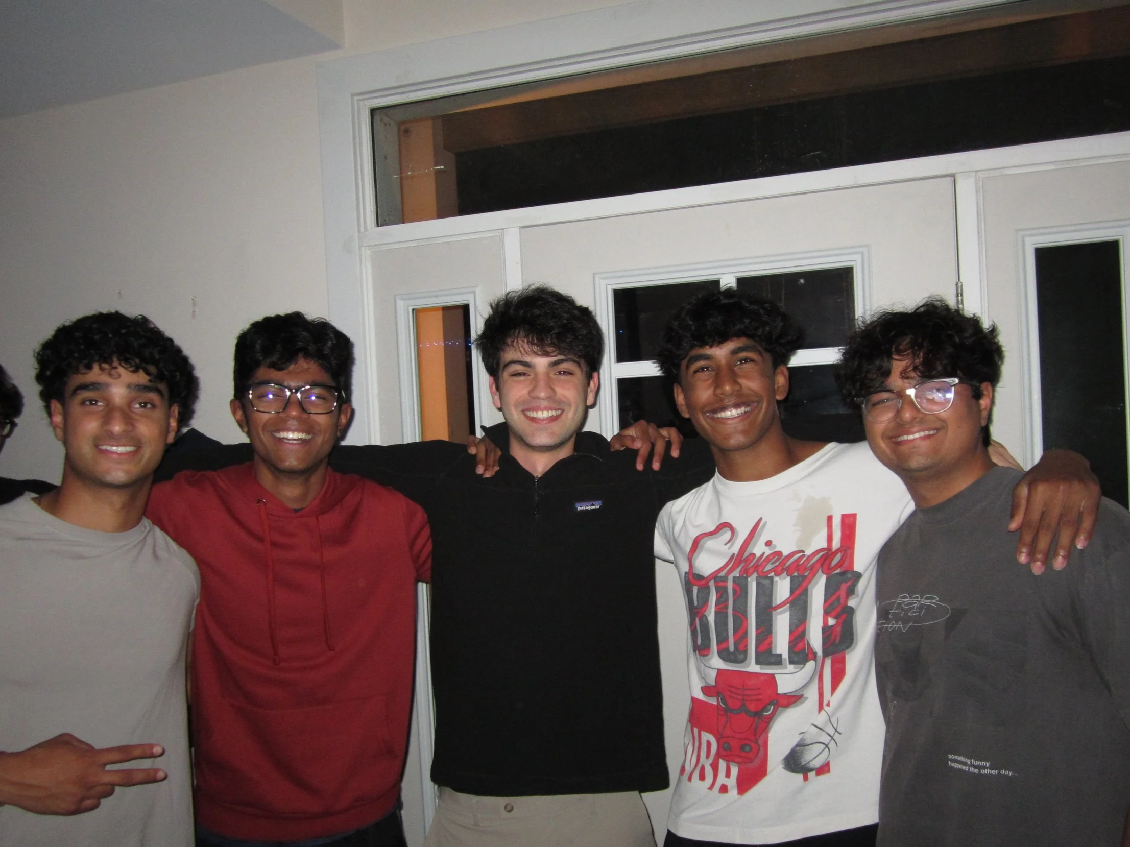 Five young men with curly hair smiling and posing with arms around each other indoors in front of a large window and white door.