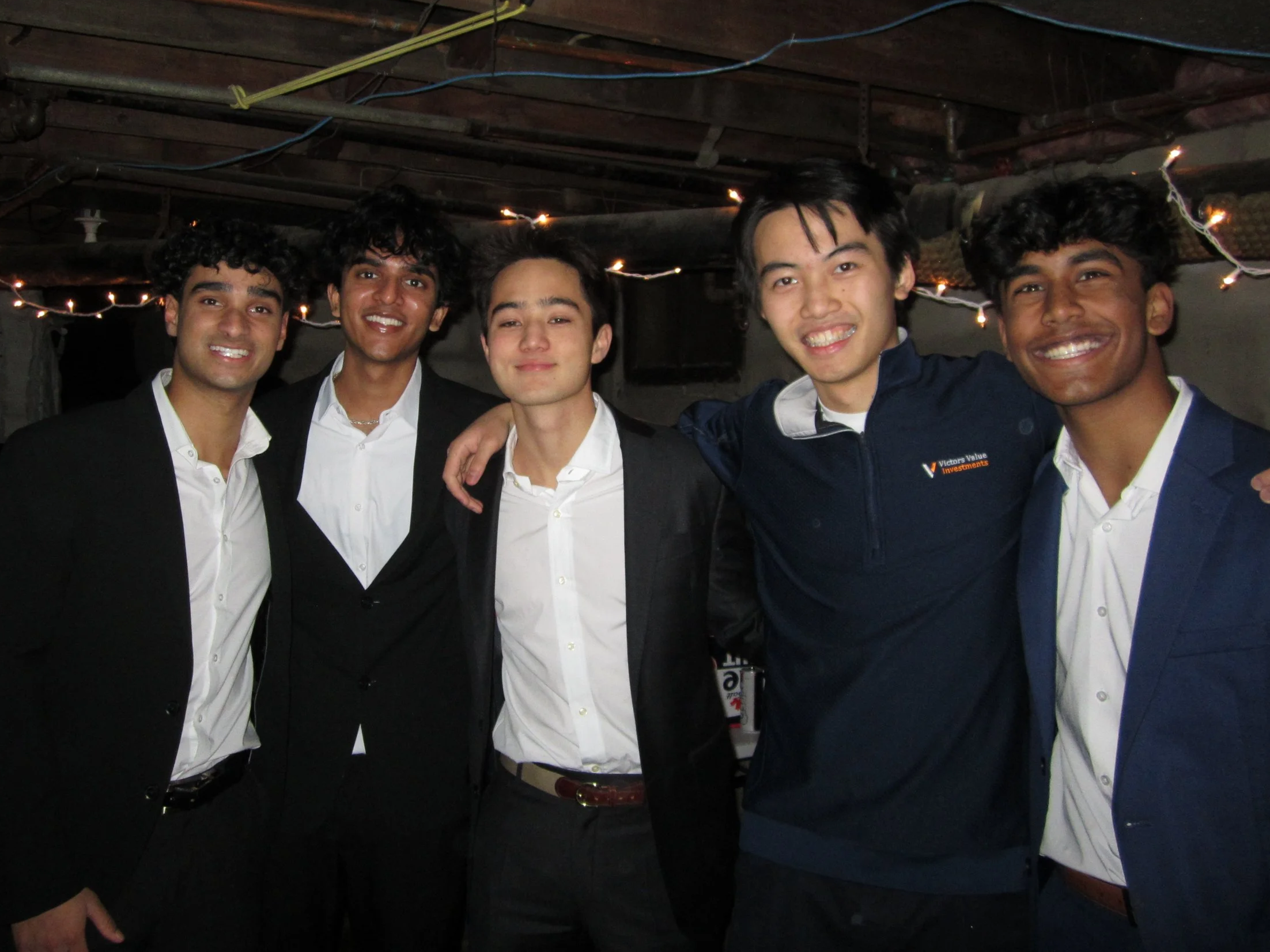 Five young men dressed in formal attire, standing close together with arms around each other, smiling indoors at a party or celebration.