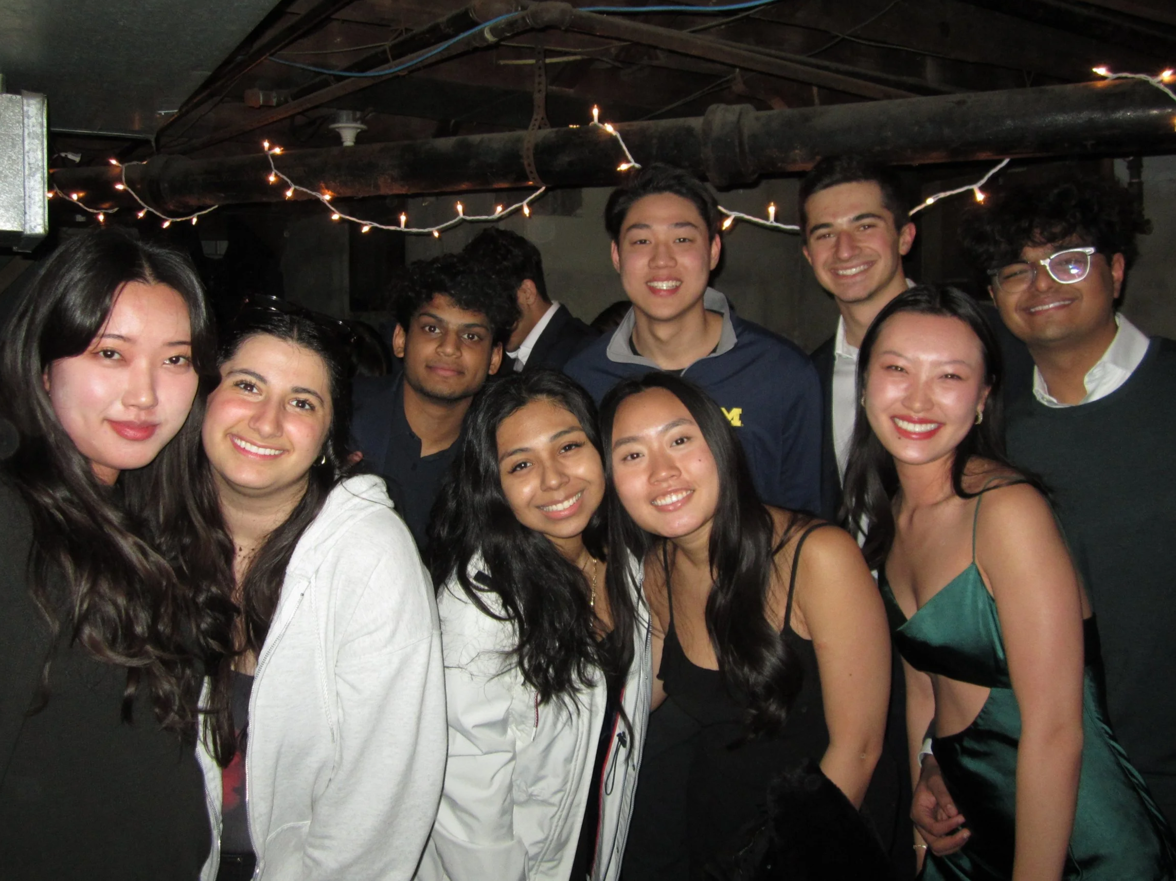 Group of ten young adults posing together at a party with string lights overhead, smiling for the camera.