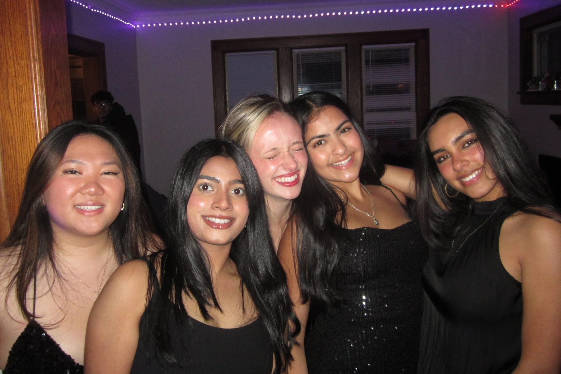 Group of five young women in black dresses smiling and posing together at a party, with some women embracing each other in a room decorated with purple and pink string lights.