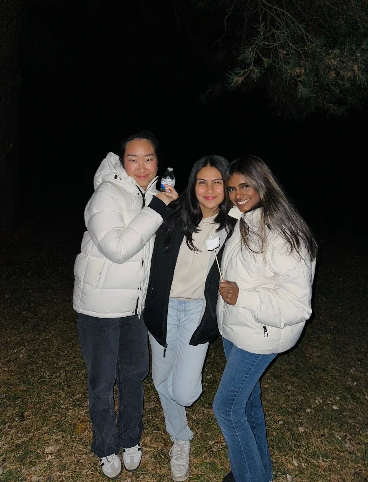 Three young women standing outdoors at night, wearing winter jackets, smiling at the camera. One woman is holding a water bottle, another is holding a marshmallow on a stick, and they are close together.