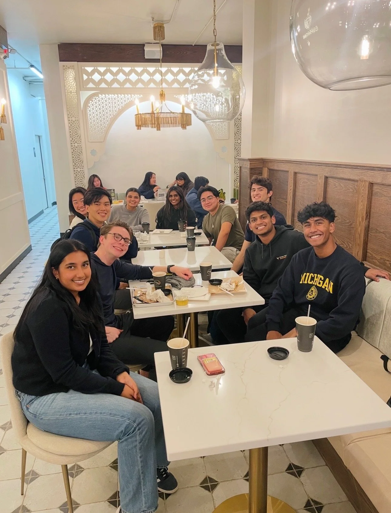 A group of young adults sitting at a long table in a restaurant, smiling and enjoying their meal.