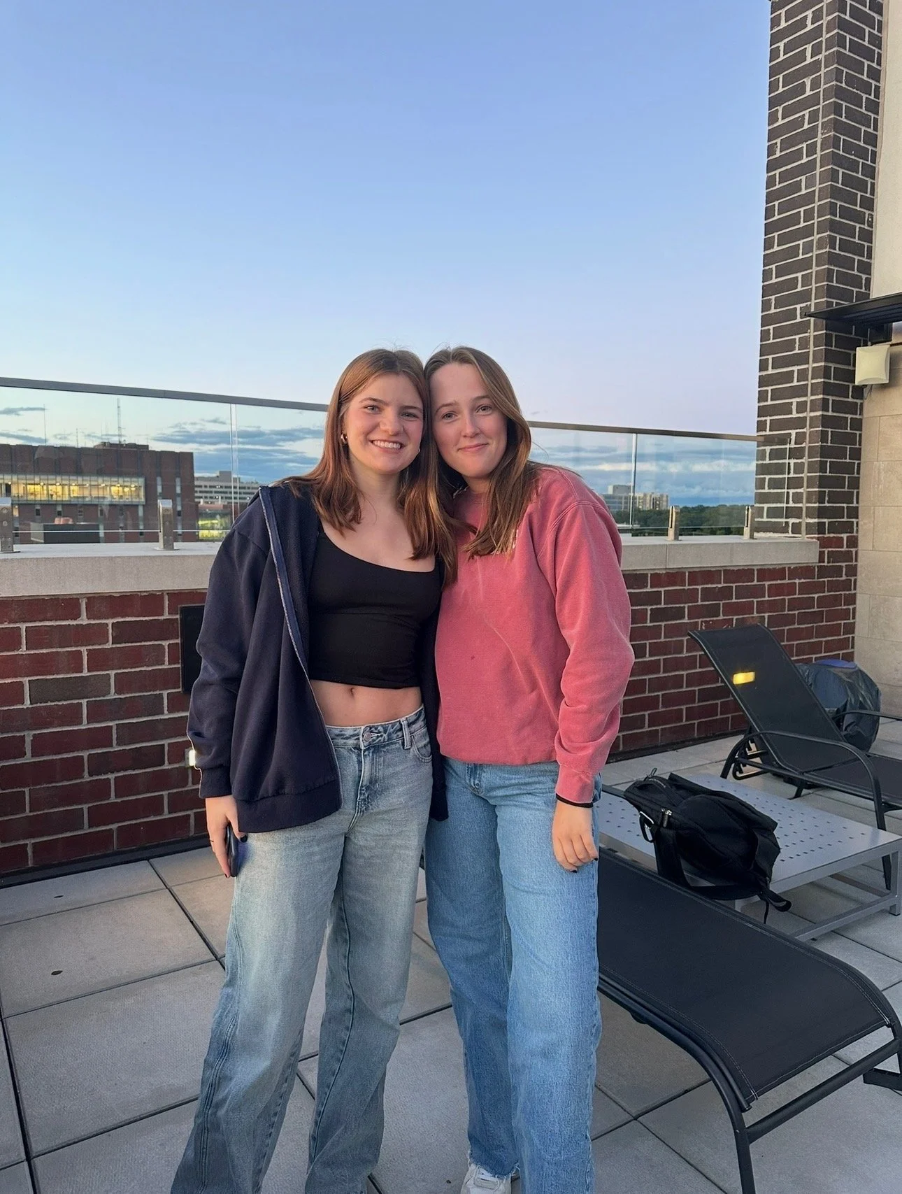 Two young women standing close together on a rooftop balcony, smiling at the camera, with city buildings and a partly cloudy sky in the background.