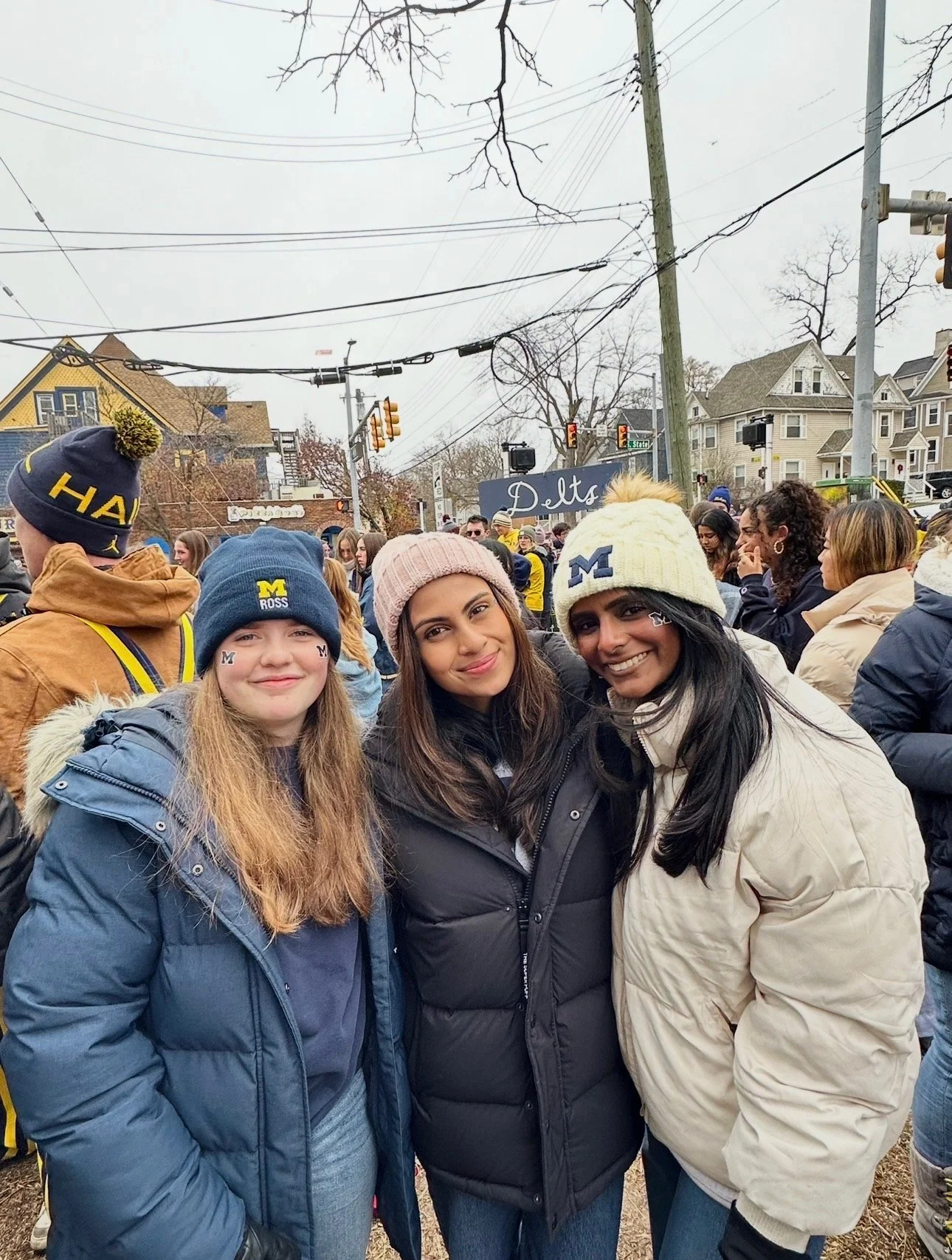 Three women standing outdoors among a crowd, wearing hats and jackets supporting the University of Michigan.