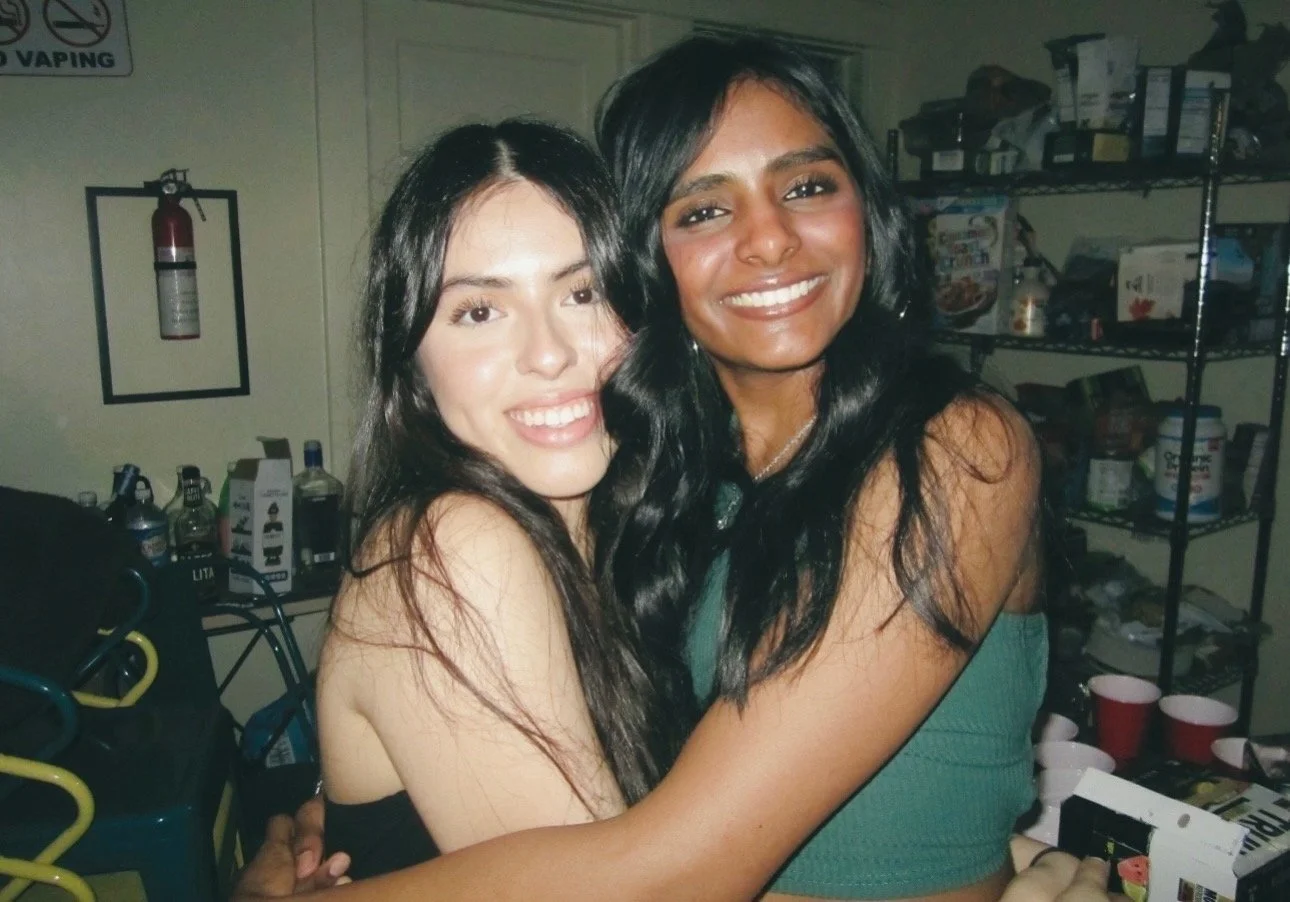 Two women smiling and hugging each other in a room with shelves stocked with food and drinks.