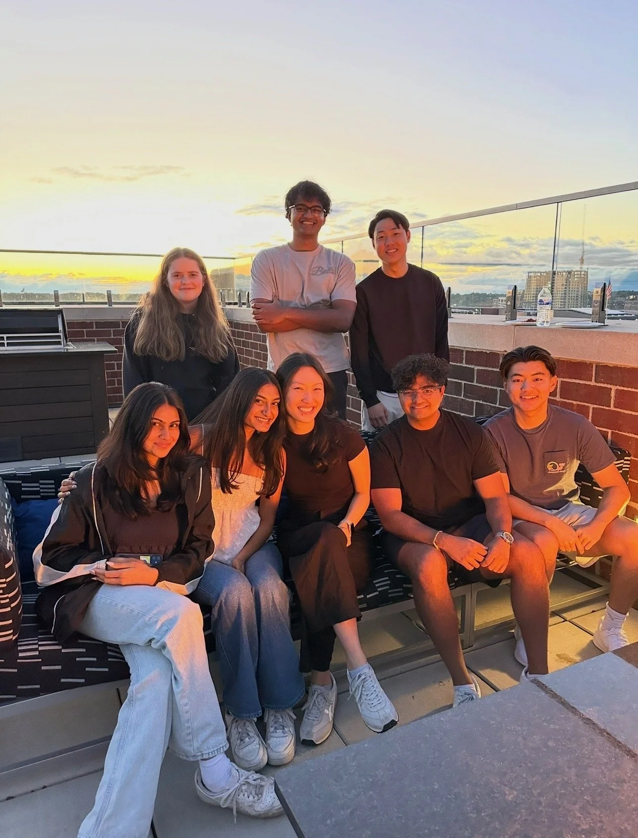 Group of nine young adults sitting and standing on a rooftop patio during sunset, smiling at the camera.