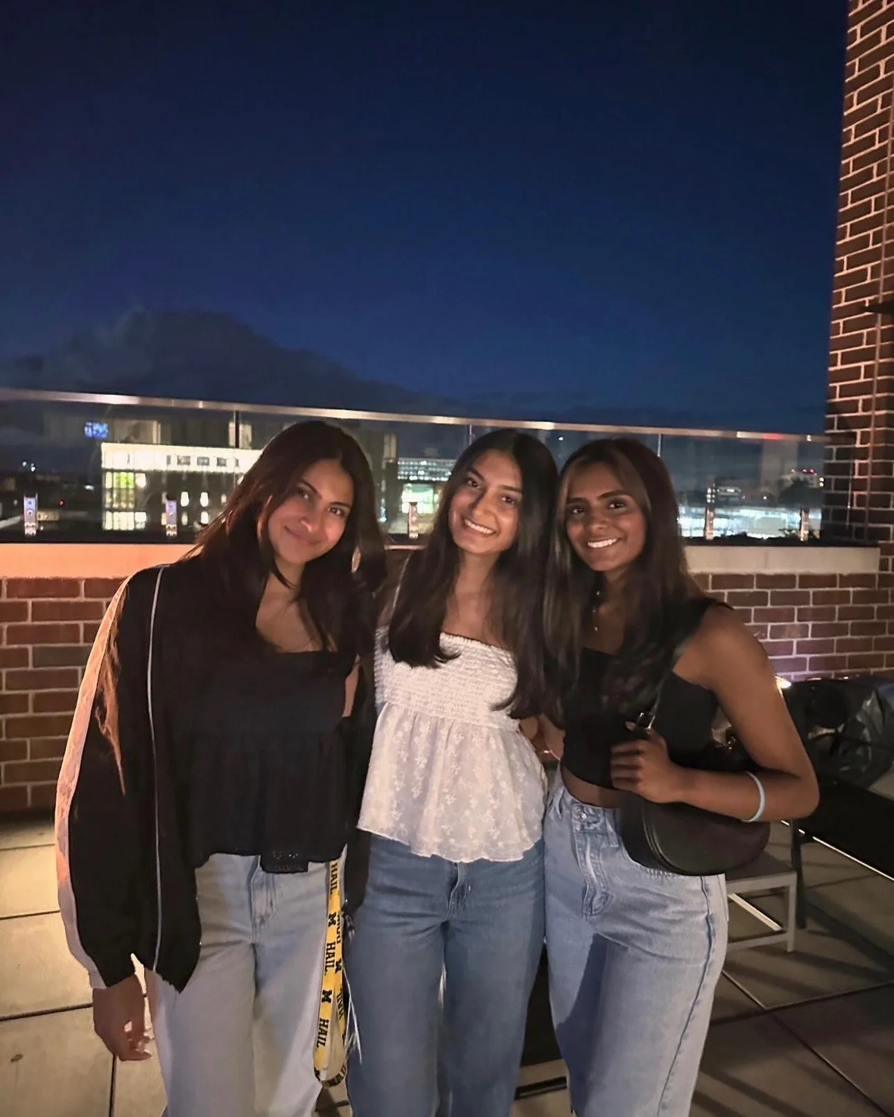 Three women smiling and standing close together on a rooftop with city lights in the background at night.