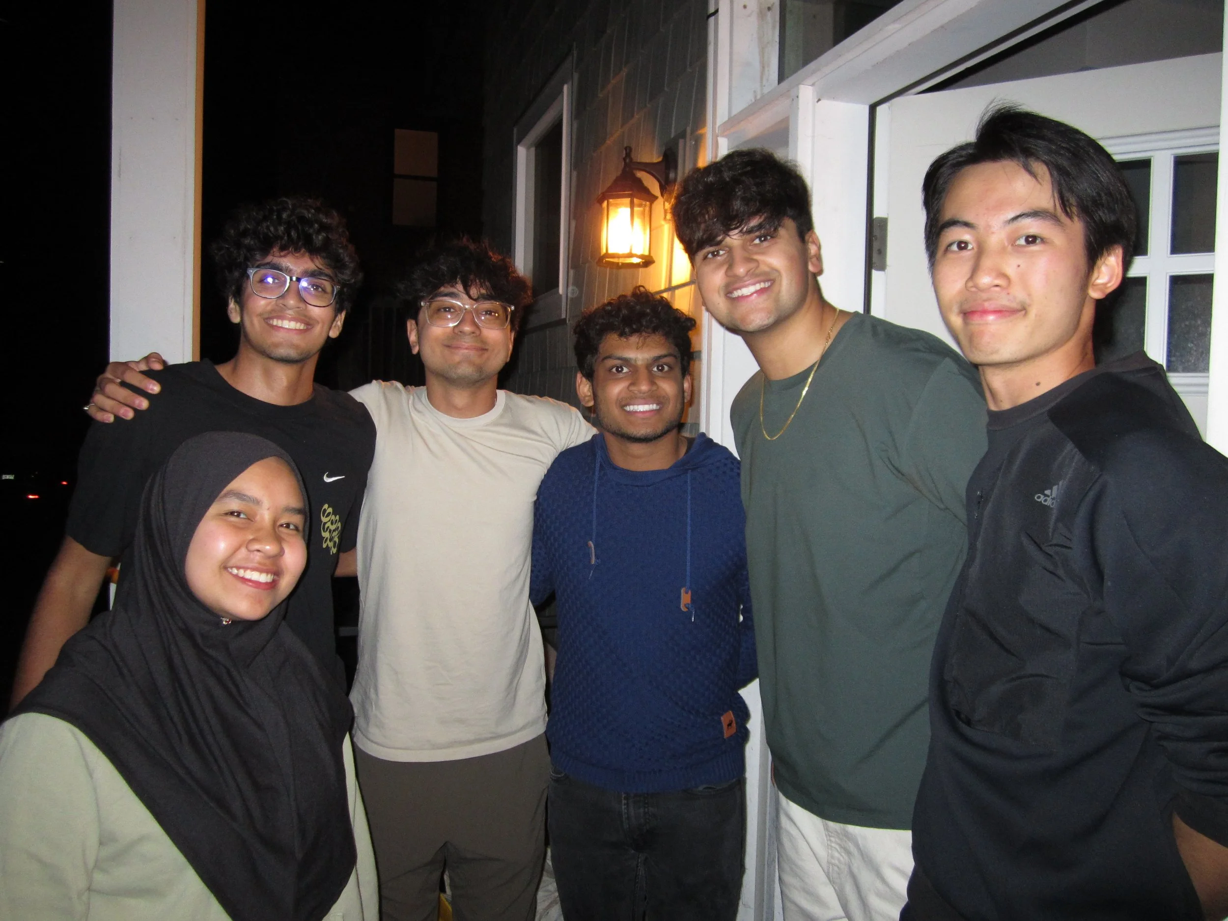 A diverse group of six young people standing close together outside at night, smiling for the camera, with a lit lantern on the wall behind them.