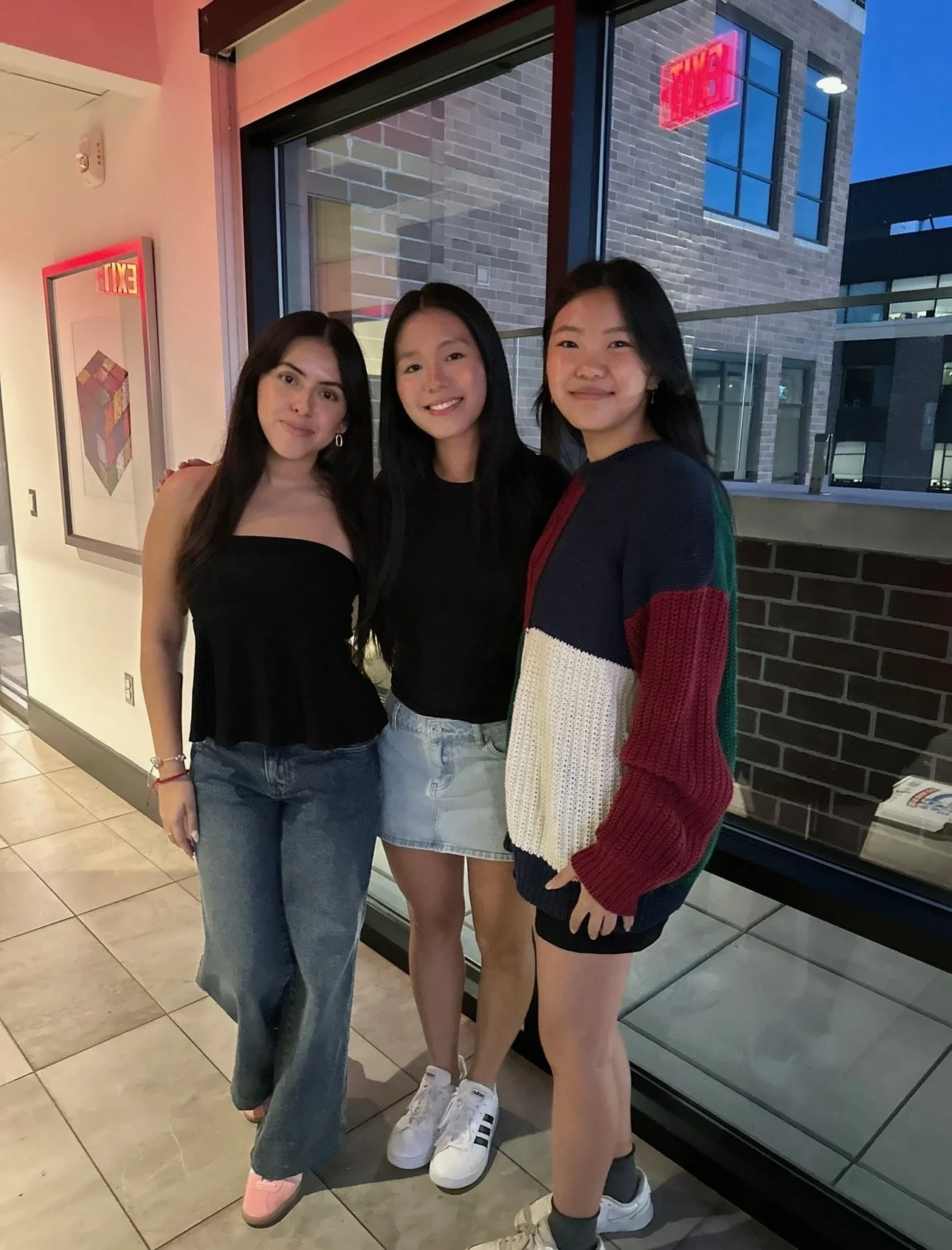Three young women standing together inside a building near a large window, smiling at the camera, with city buildings visible outside.