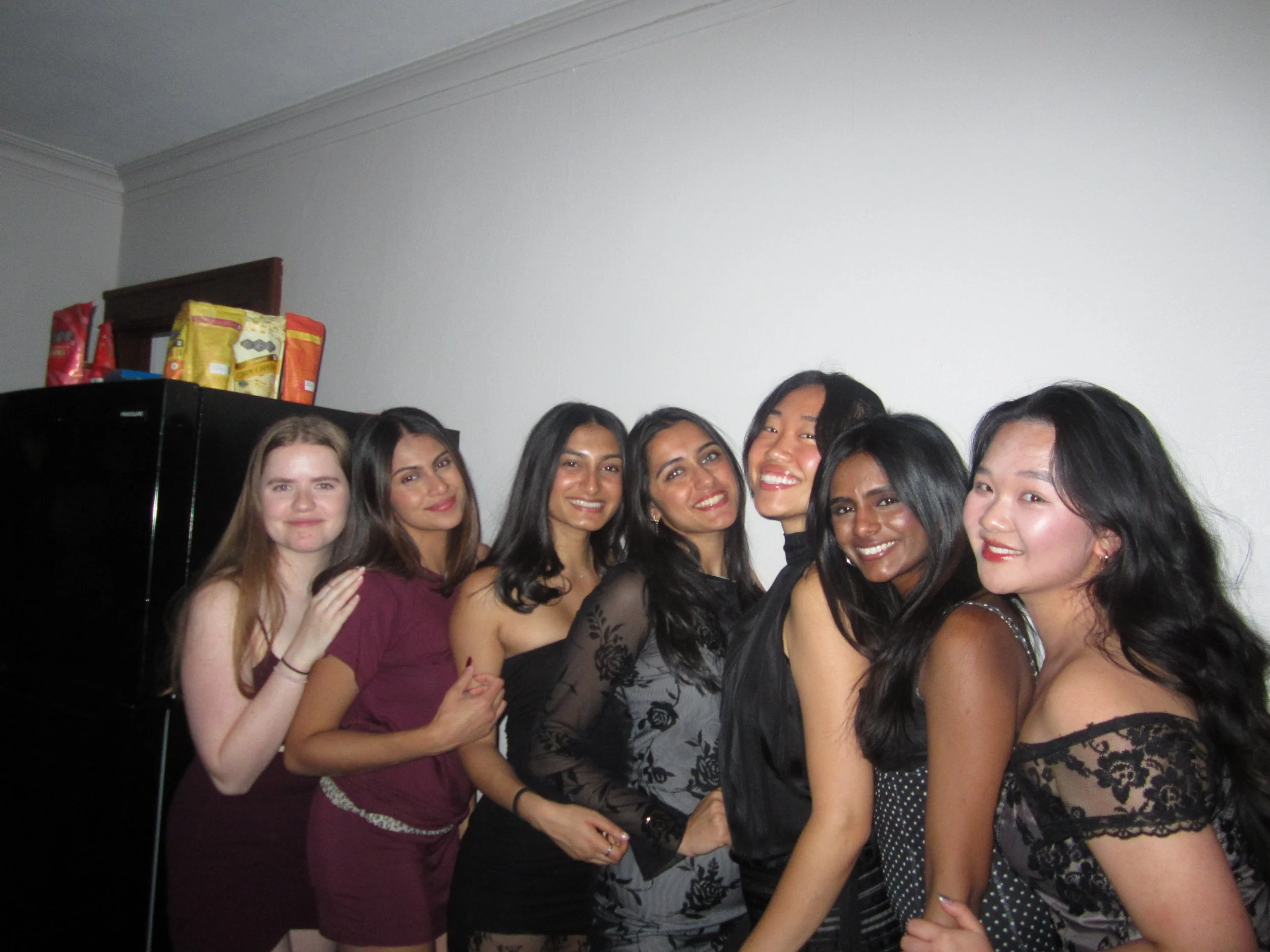 Group of seven women smiling and posing together indoors, with a refrigerator and pantry in the background.