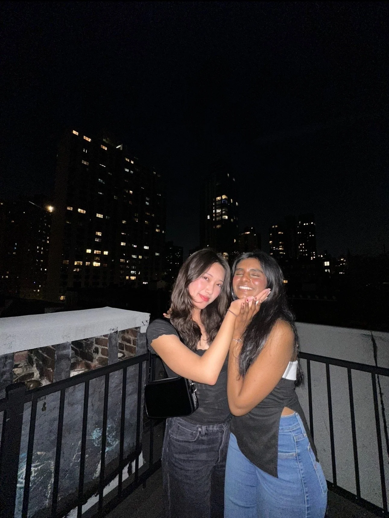 Two women smiling and holding hands on a rooftop at night, with city buildings illuminated in the background.
