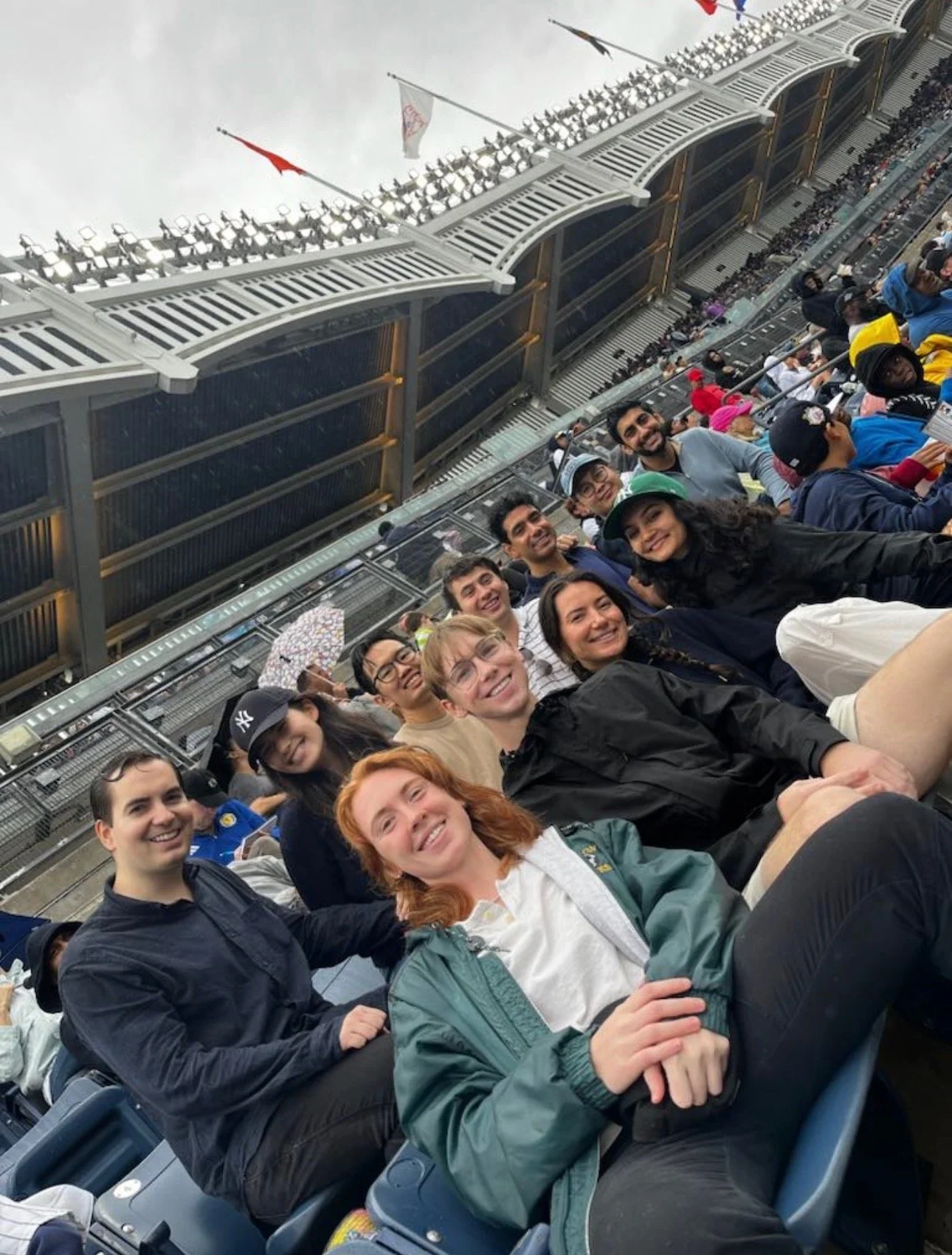 A group of people sitting in stadium seats at a sporting event, smiling at the camera under an overcast sky.