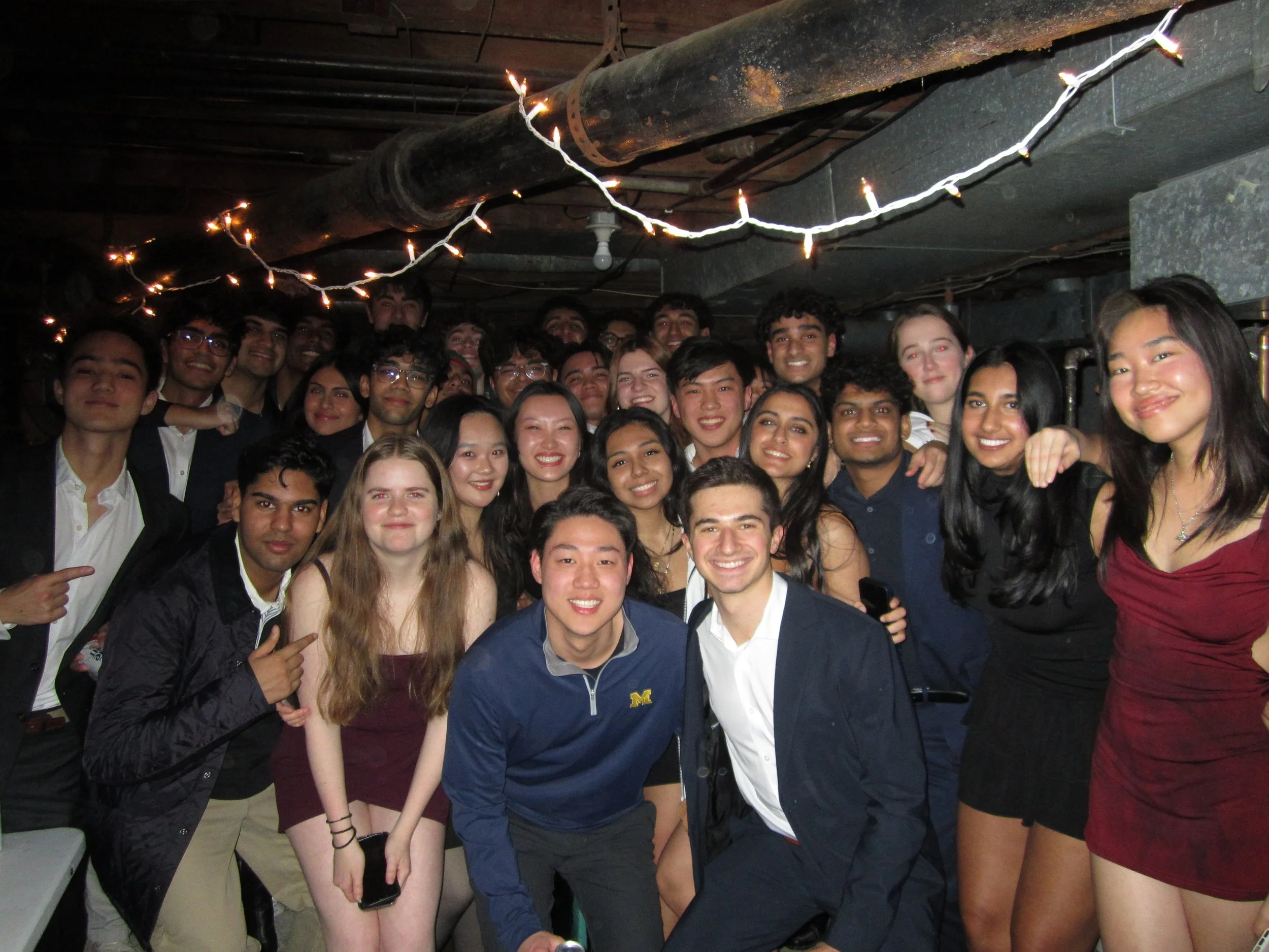 Group of diverse young people dressed in semi-formal attire, smiling at a party or celebration, under string lights in an industrial-style space.