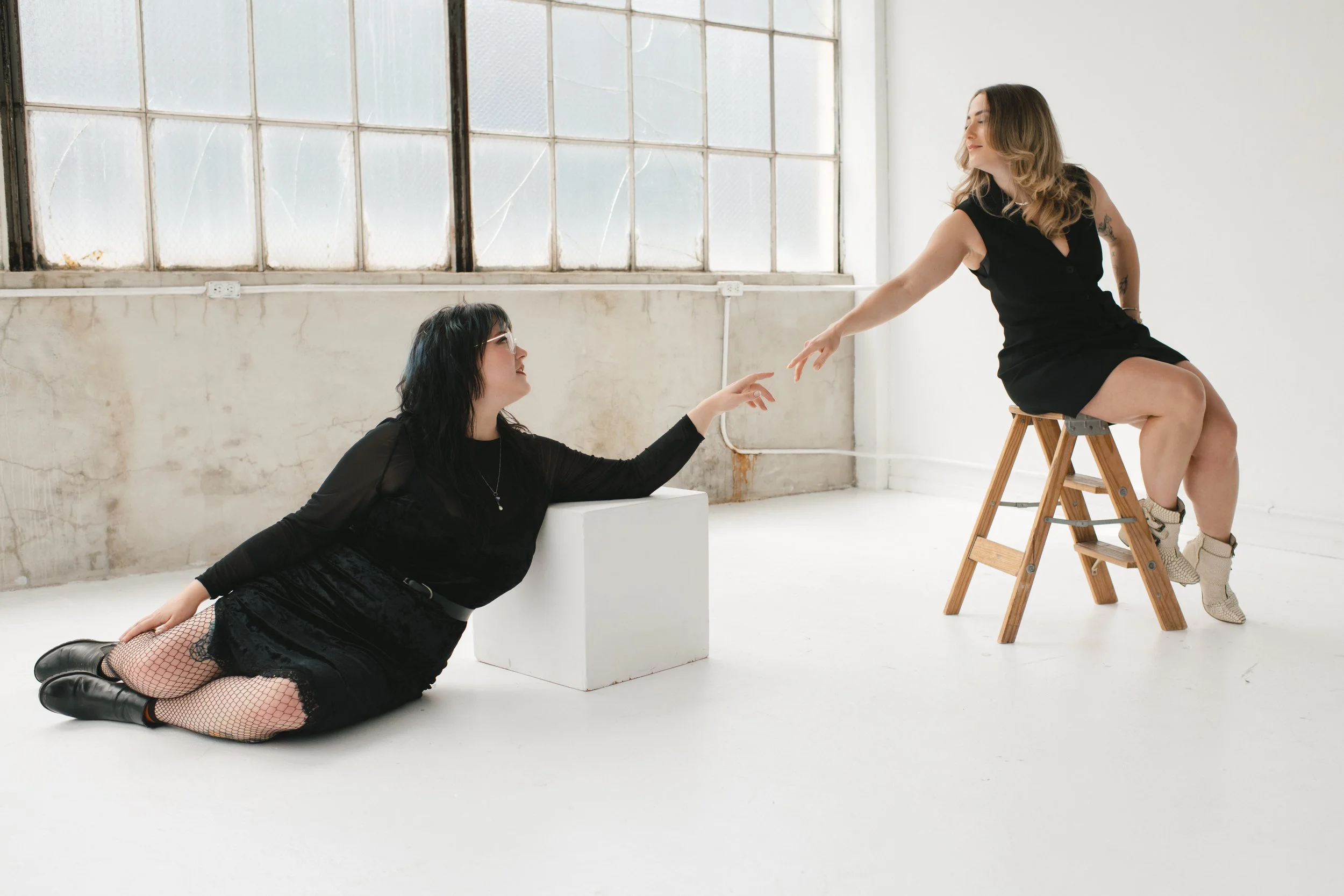 Two women in black outfits, one sitting on a stool and the other on the floor, reaching towards each other in a minimalist studio with large windows.
