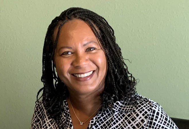 Smiling, Yvette Ward is wearing a patterned black and white blouse, sitting against a light green wall.
