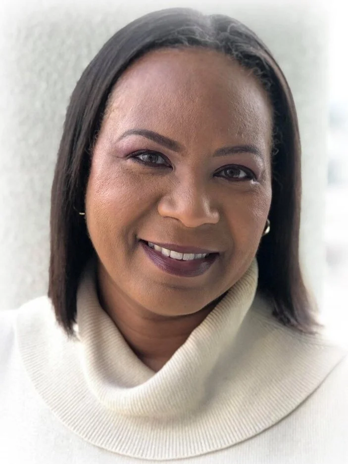 Close-up of Yvette Ward, smiling with shoulder-length dark hair, wearing a cream turtleneck sweater, standing against a light background.