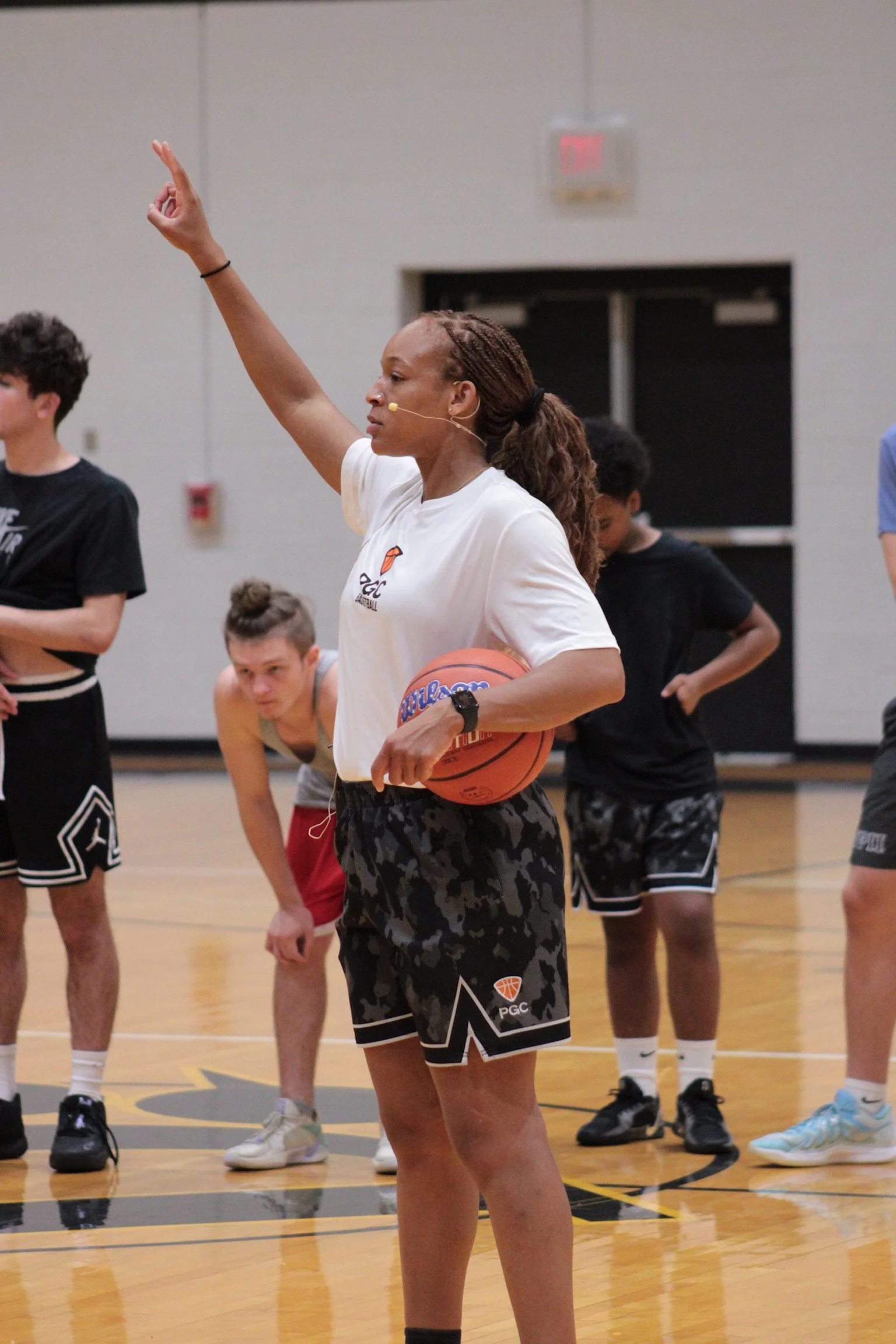 Taneka PGC Director in Action, right hand in the air, basketball under left arm at her waist, communicating with the athletes through her head microphone.