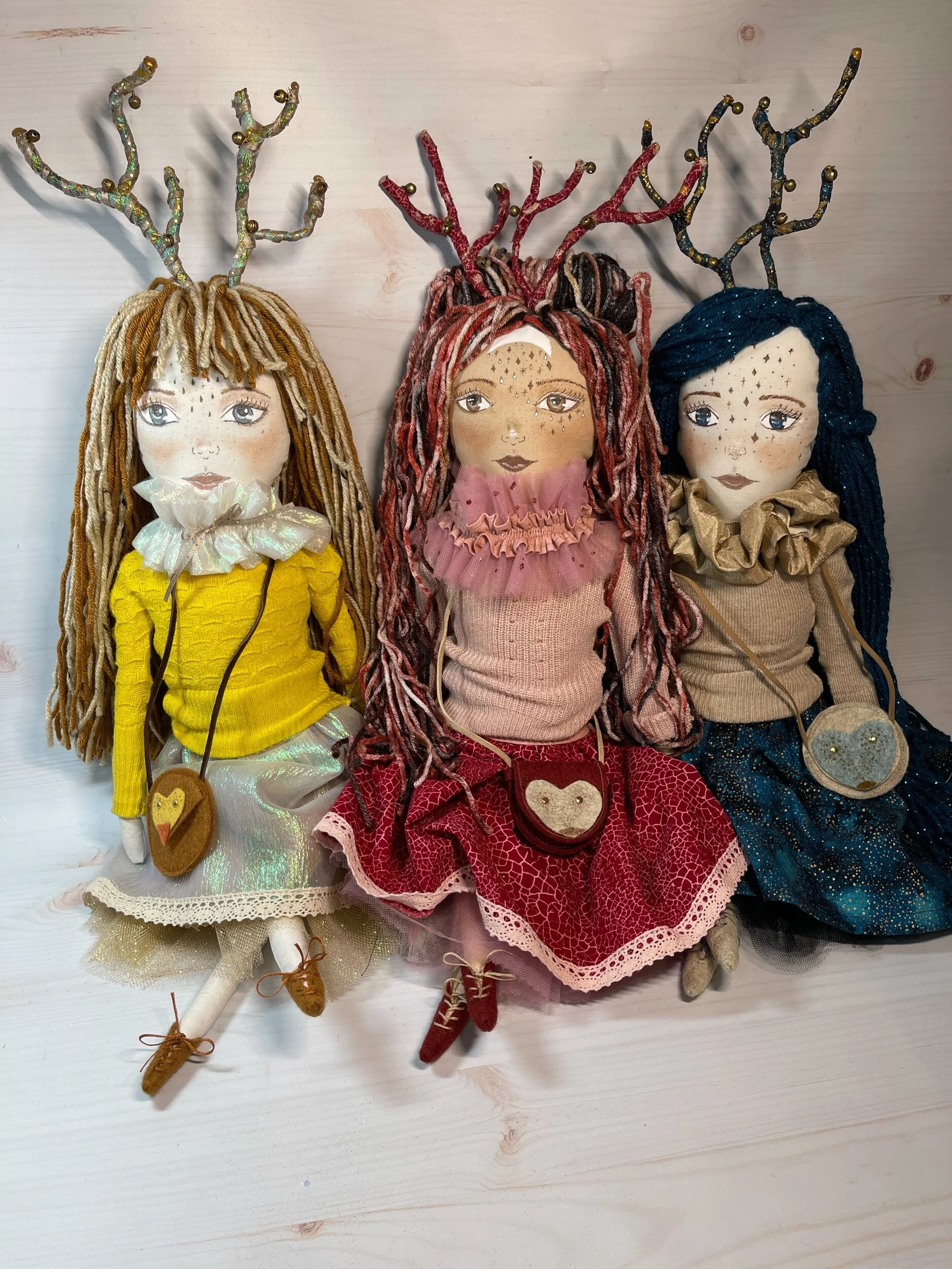 Three handmade dolls with antler-like decorations, unique hairstyles, and colorful clothing sitting against a wooden background.